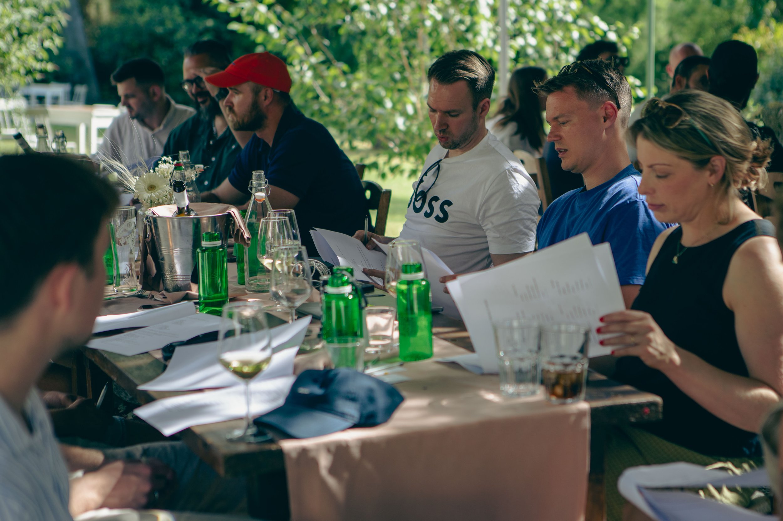 People sitting at a long outdoor table during daytime, reading menus, with drinks and flowers on the table, surrounded by trees.