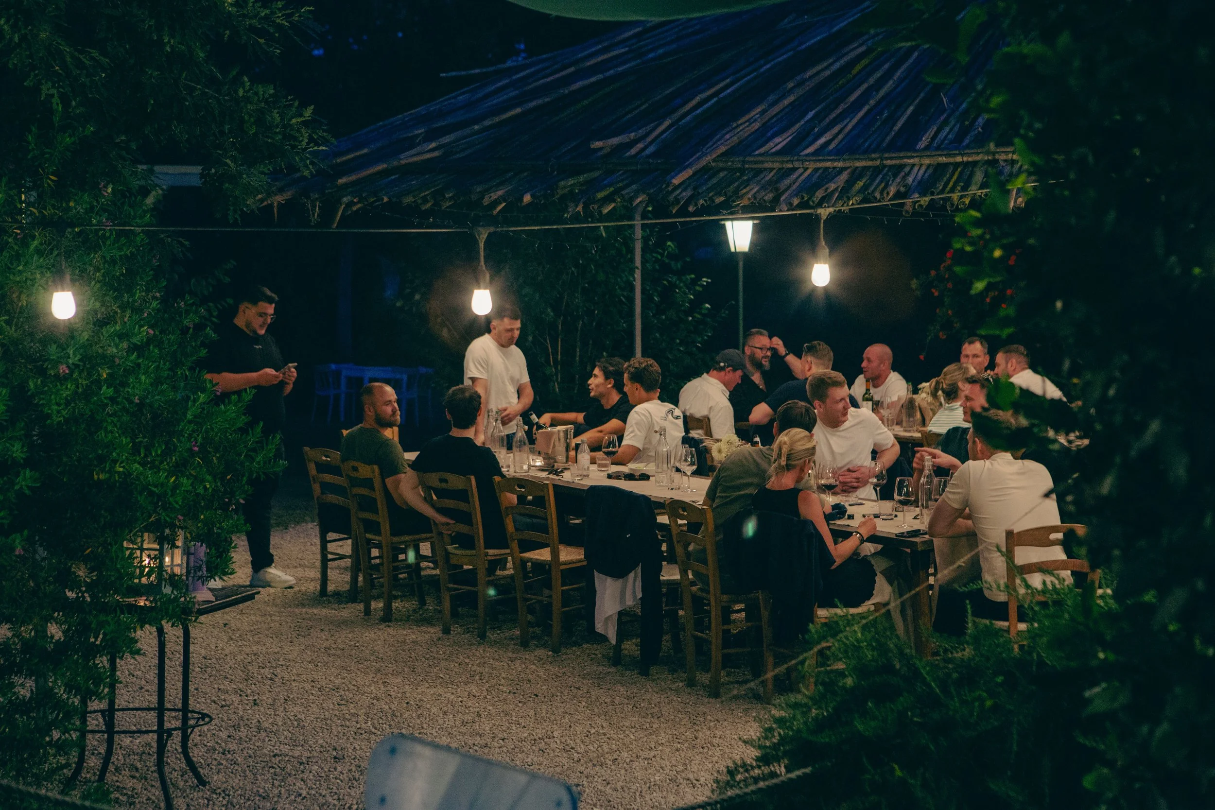 Night scene of an outdoor dinner party with a large group of people seated around a long table, illuminated by hanging lights, with a rustic roof overhead and greenery surrounding the area.