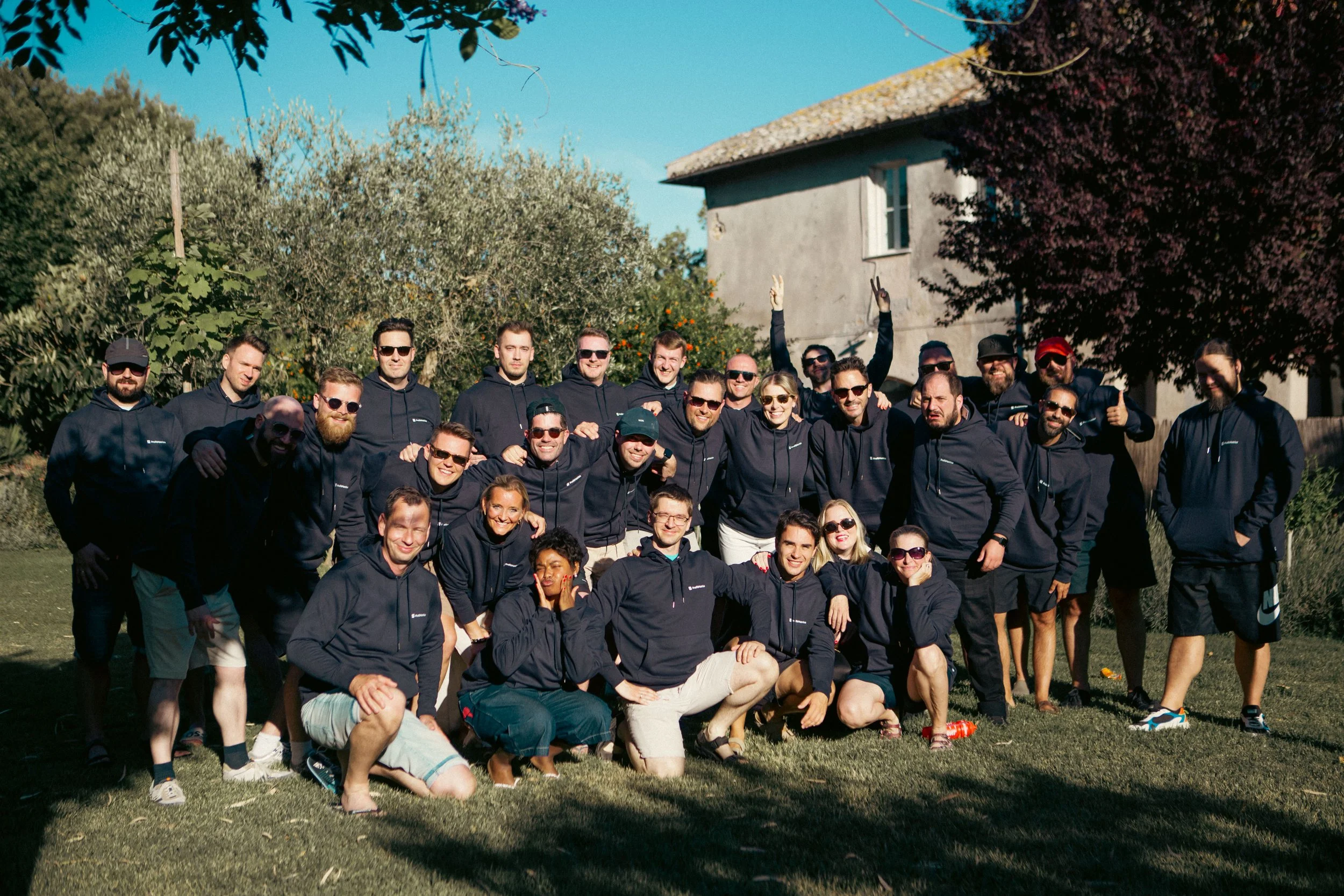 A large group of young adults posing outdoors in front of trees and a house on a sunny day. They are smiling, some wearing sunglasses, and dressed in dark hoodies and casual clothing.