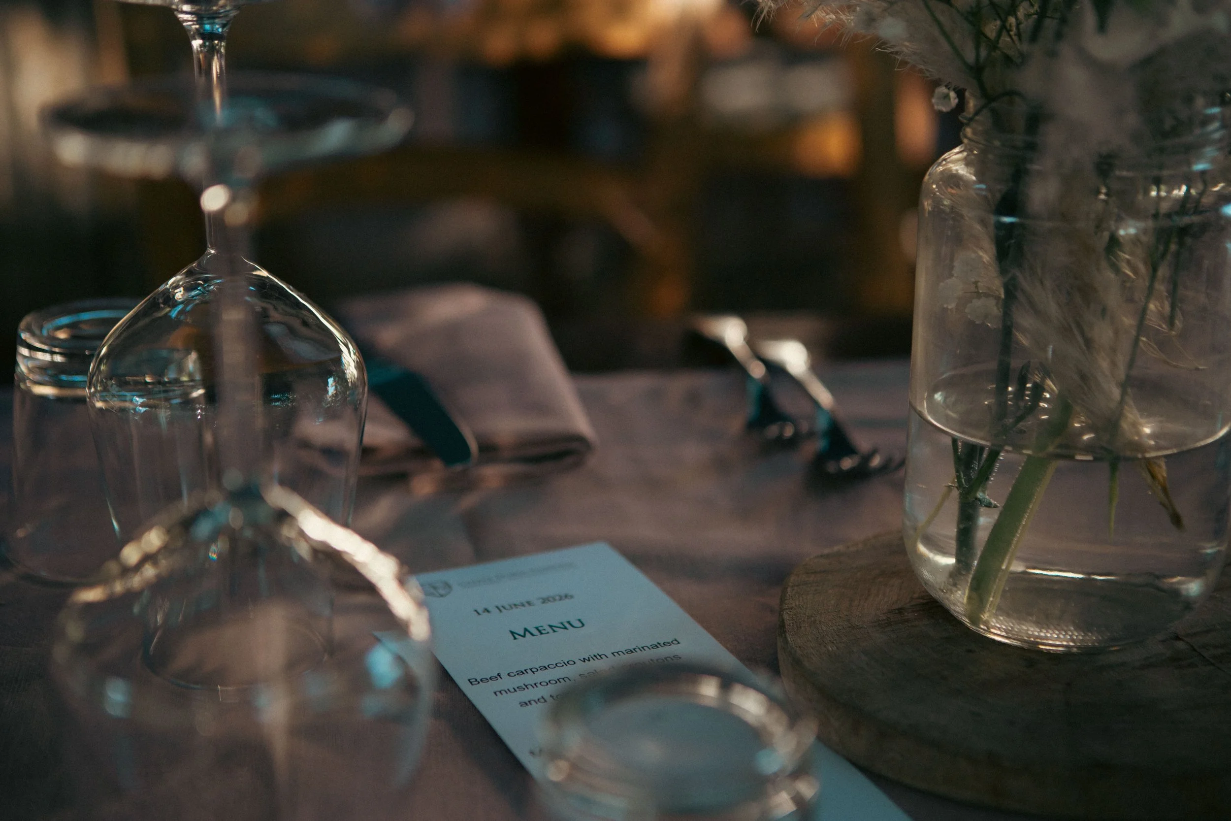 A table setting with a wine glass, a glass vase holding flowers, some scattered cutlery, and a menu card, with blurred background lighting.