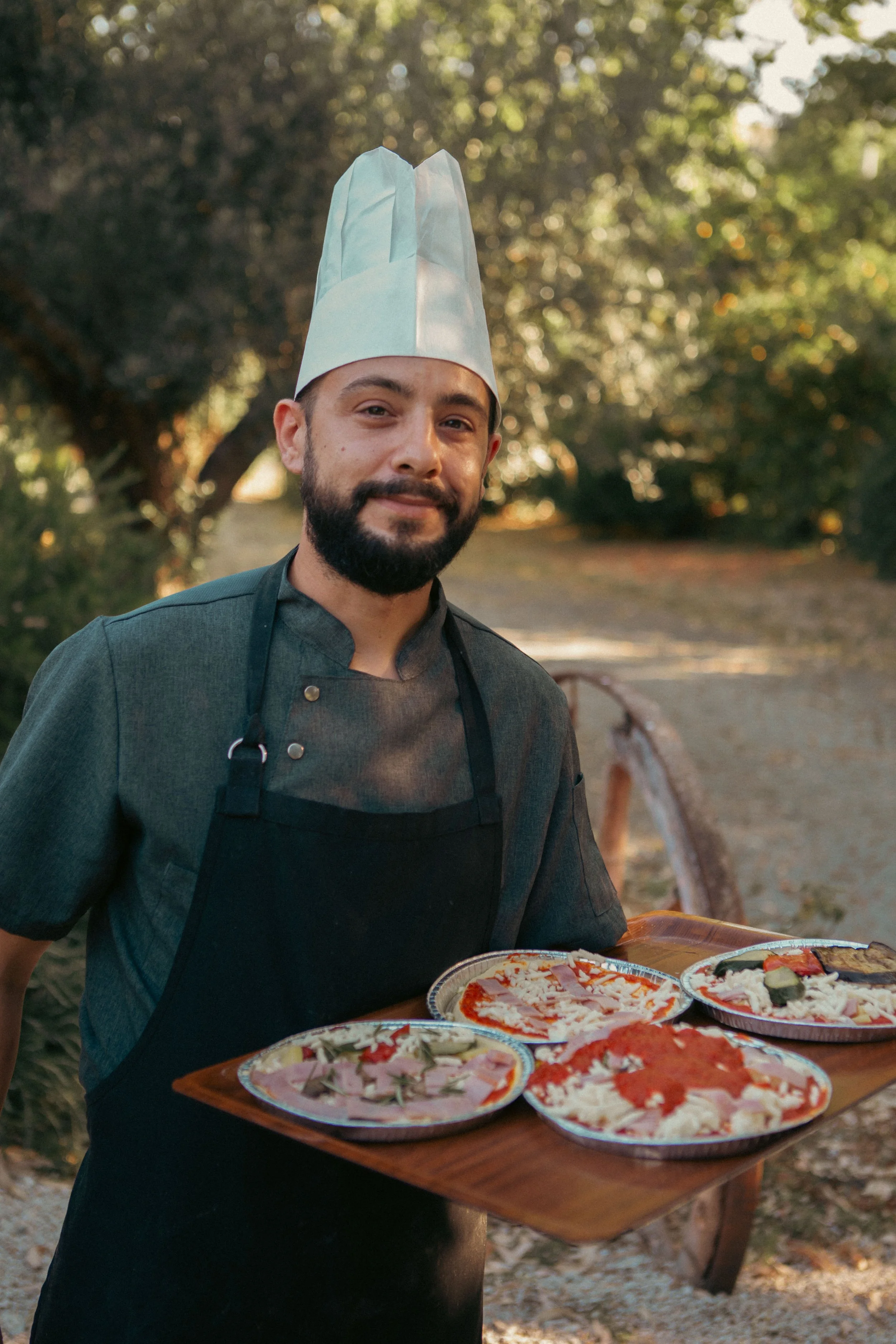 A man wearing a white chef hat and black apron holding a wooden tray with various pizzas outdoors during daytime, with a background of trees and sunlight.