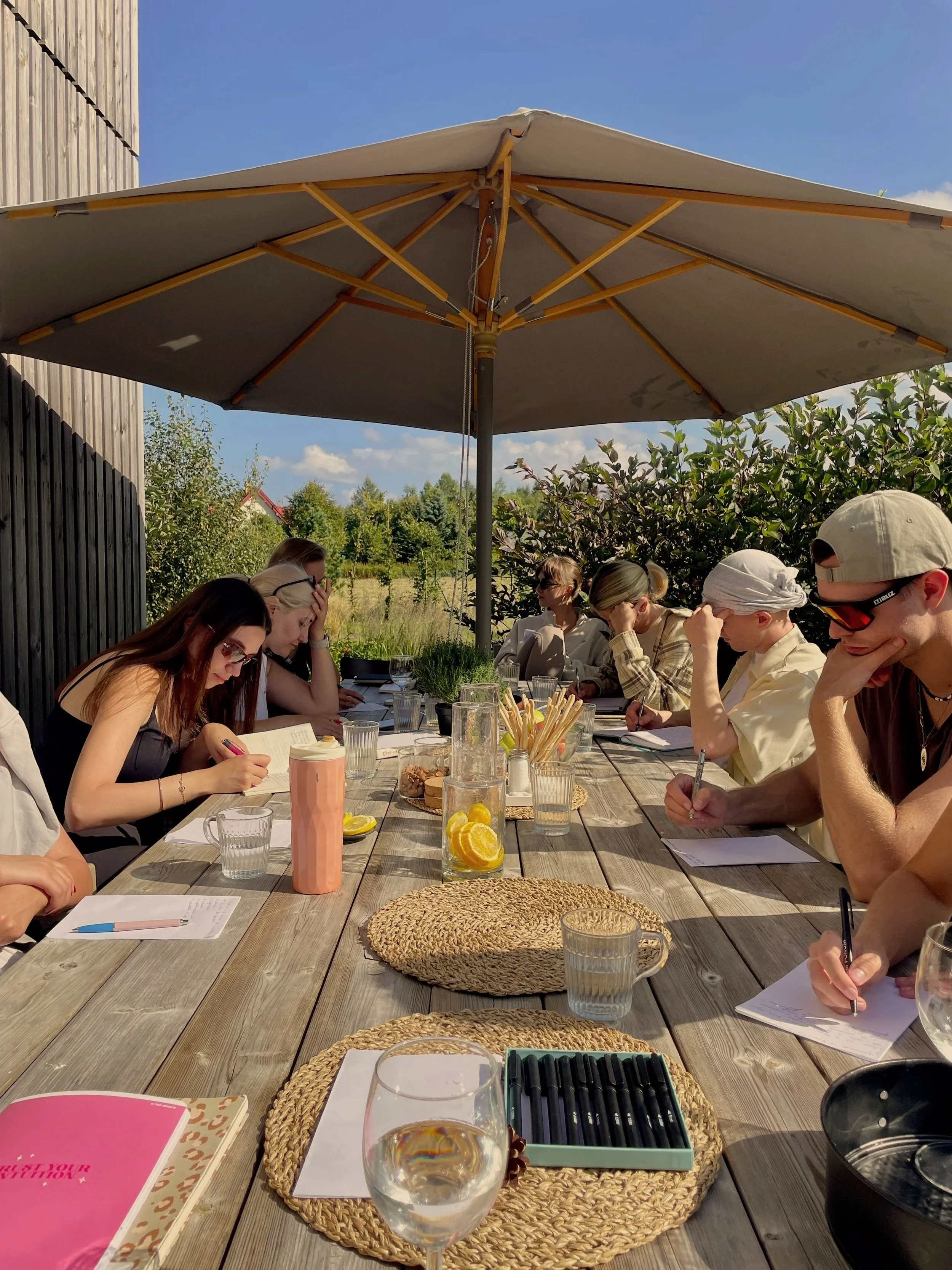 People sitting around a long wooden outdoor table under a large beige umbrella, with glasses of water, notebooks, and various items on the table, in a garden setting with trees and a clear blue sky.