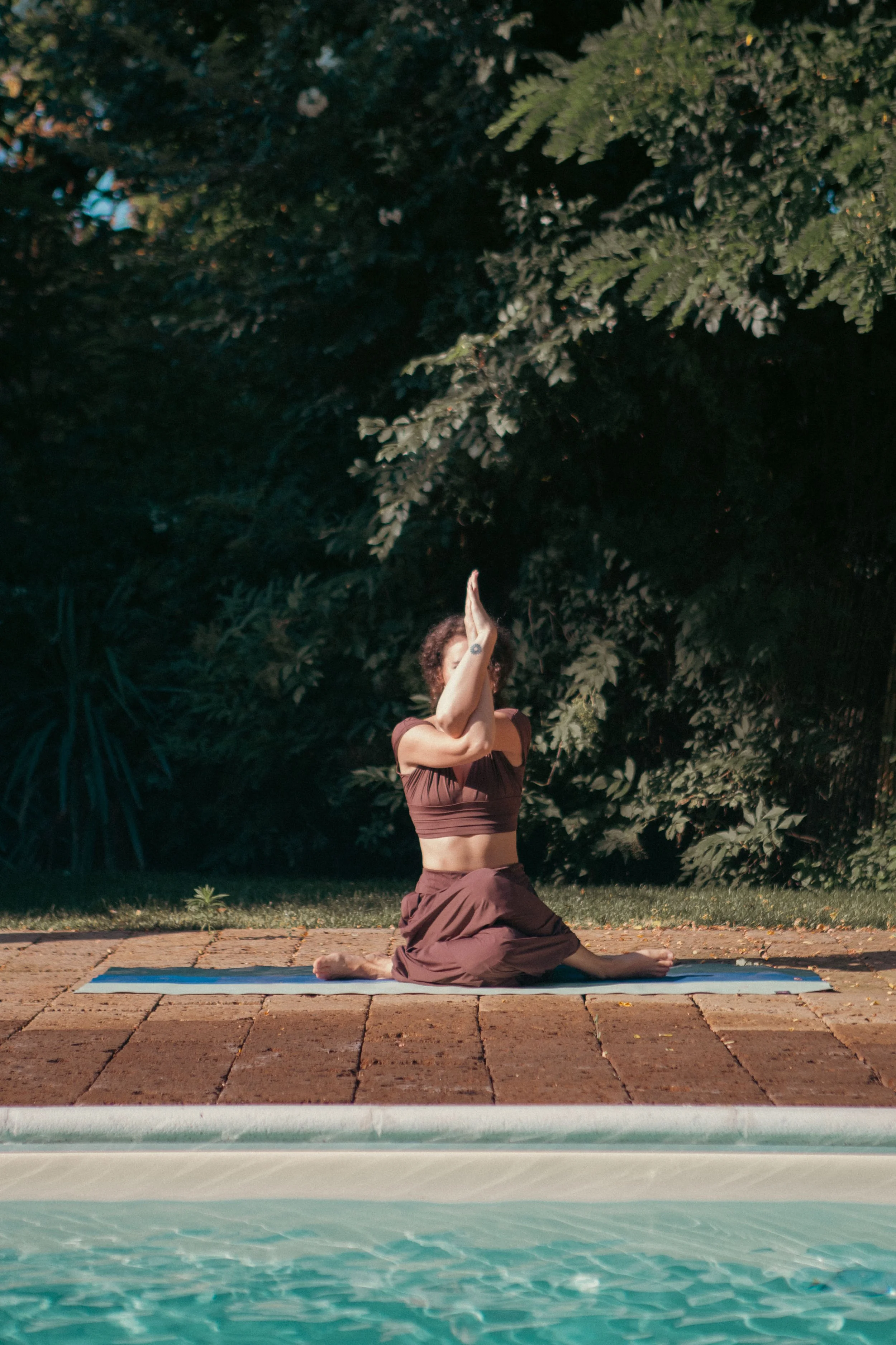A woman practicing yoga outdoors at night, kneeling on a yoga mat with her hands in prayer above her head, surrounded by lush green trees.