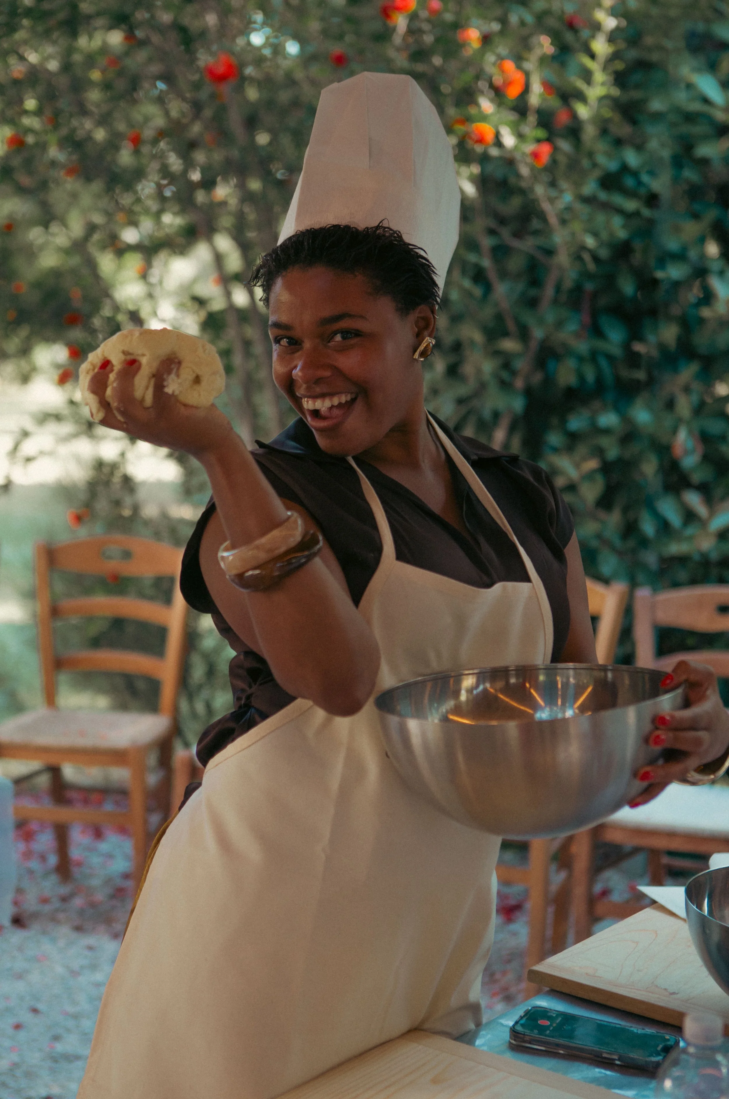 A smiling woman wearing a black shirt, beige apron, and a paper chef's hat, holding a dough or bread in her right hand and a stainless steel mixing bowl in her left hand. She is standing outdoors with trees and orange flowers in the background.