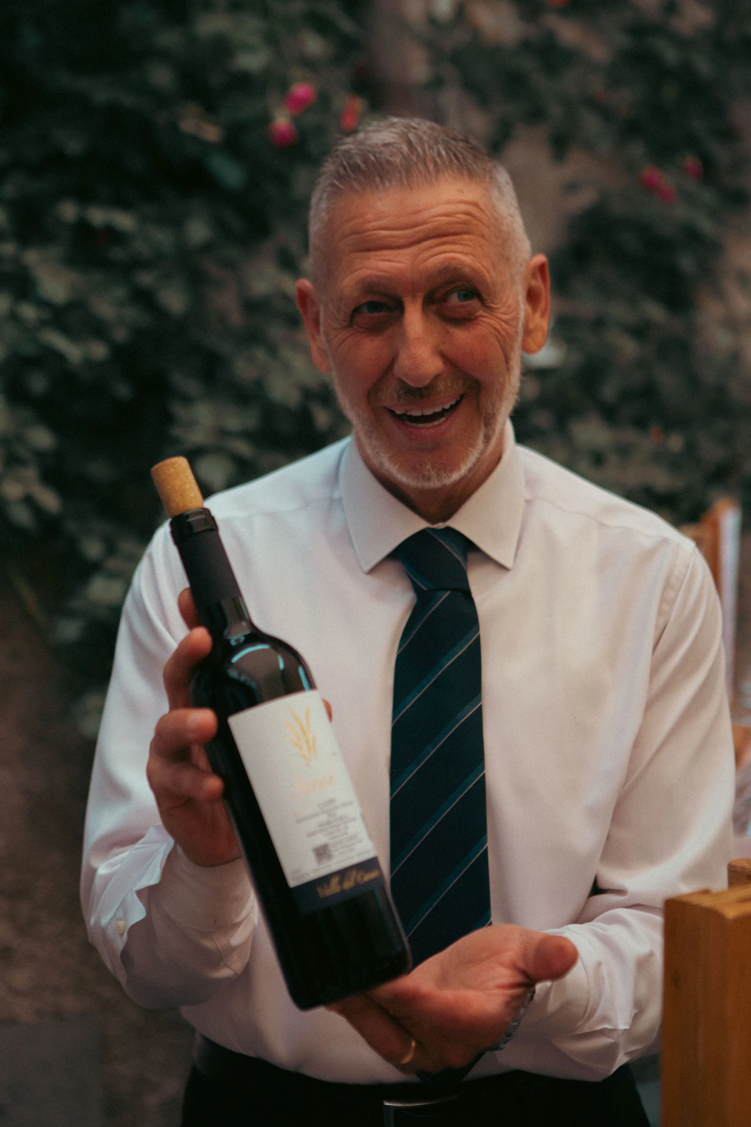 A cheerful older man with gray hair and a beard, wearing a white shirt and a dark striped tie, is holding a bottle of red wine with a white label. The background is blurred, showing green foliage and some pink flowers.