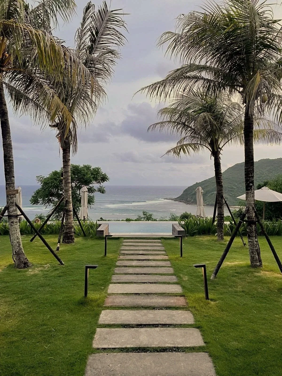 Pathway leading to a swimming pool overlooking the ocean, with palm trees and umbrellas on a cloudy day.