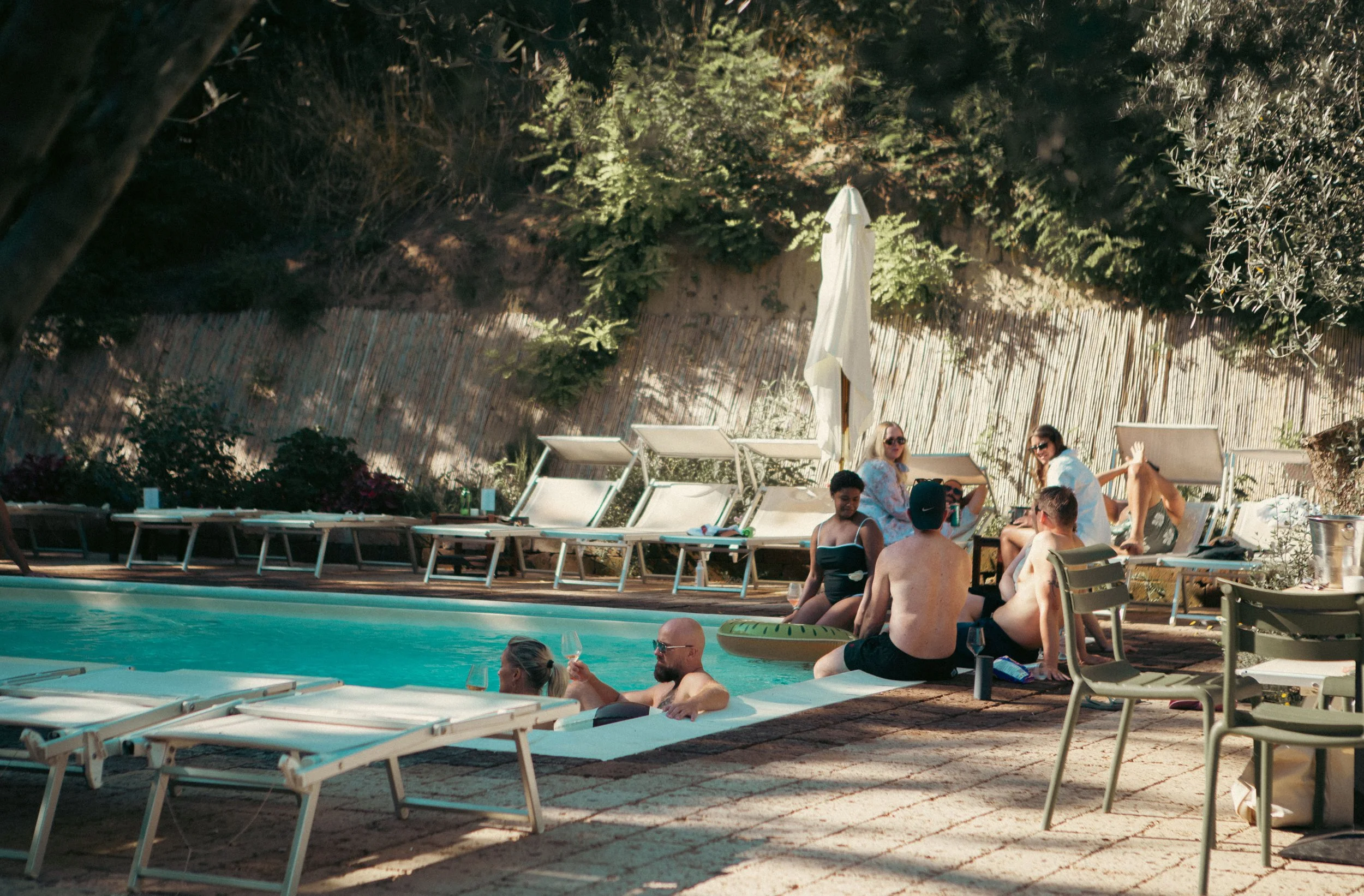 People relaxing and socializing by a swimming pool at a backyard or outdoor lounge area during daytime, with lounge chairs, a large white umbrella, and a tall wooden fence in the background.