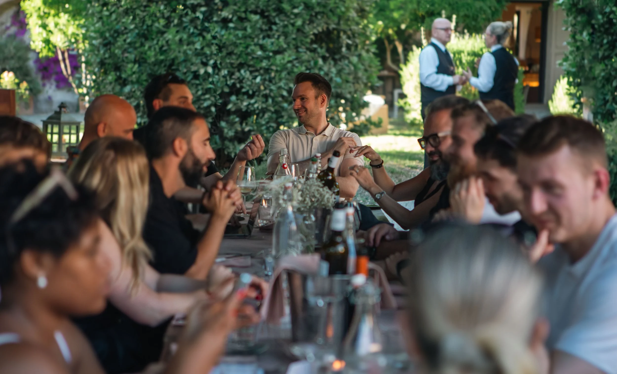 People gathered around a long table outdoors enjoying a meal and conversation, with lush greenery and trees in the background.