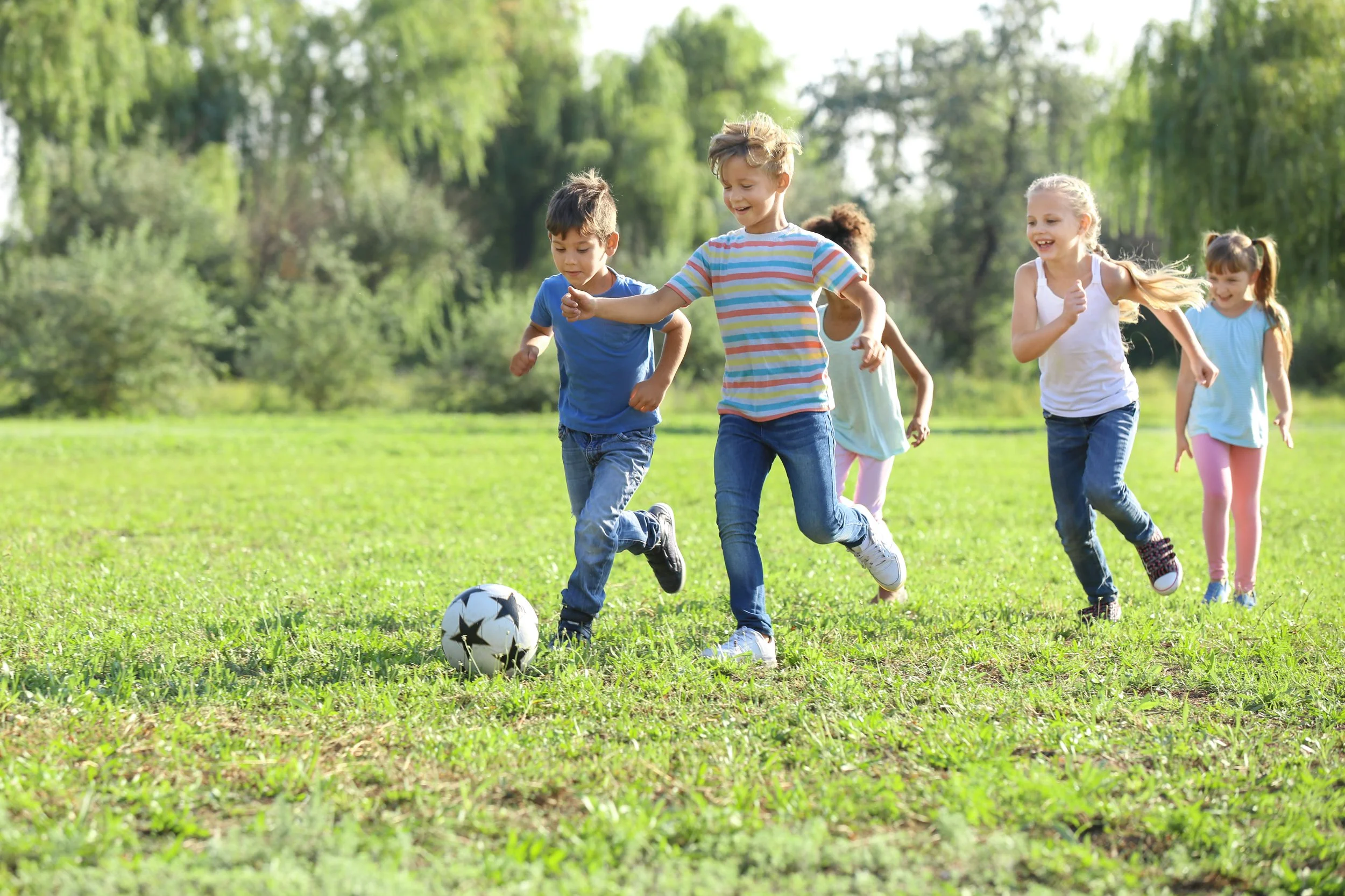 Group of children playing soccer on a grassy field in a park on a sunny day.