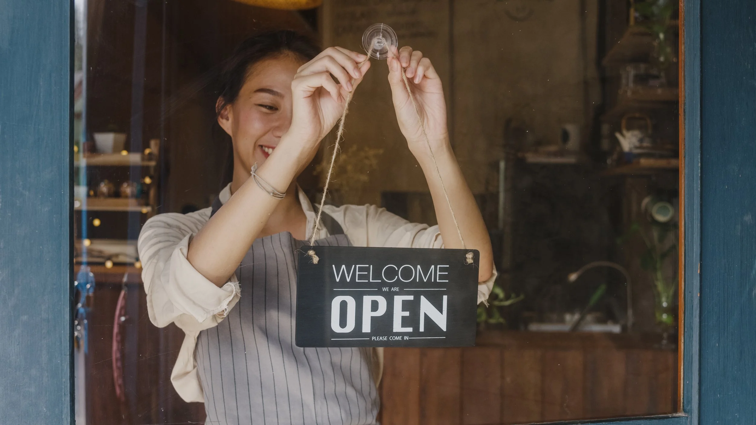Smiling woman hanging a 'Welcome, we are open' sign on a cafe door window.