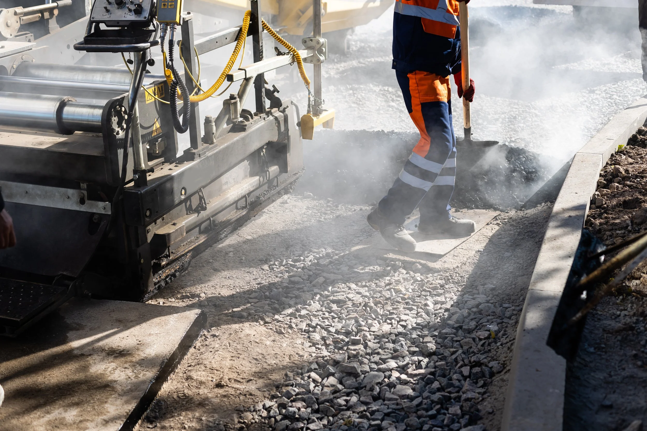 Worker in safety gear operating machinery that is laying down asphalt on a road, with steam or smoke rising from the material.