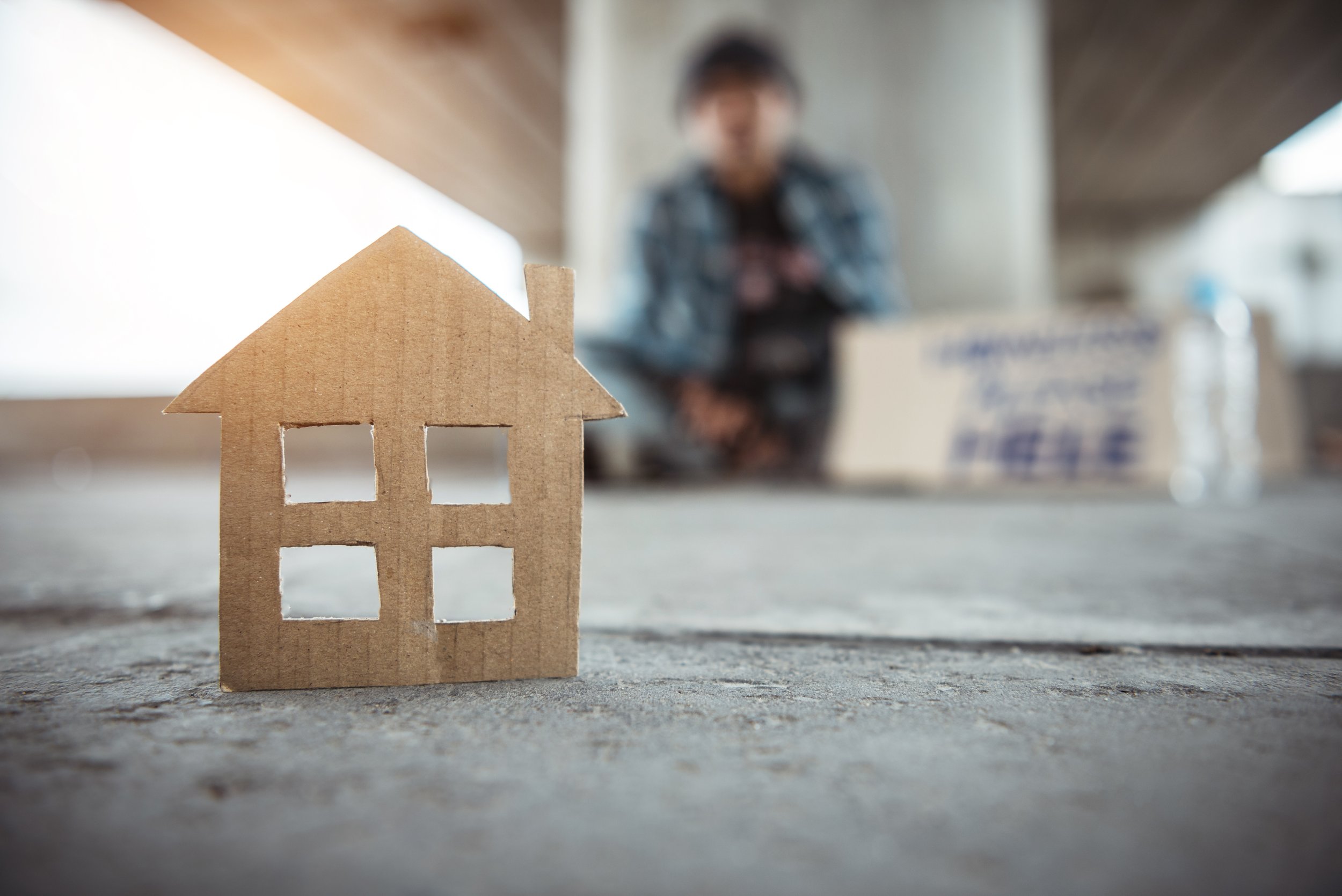 A small cardboard house model with four window cutouts on a concrete surface, with a blurred person and a sign in the background.