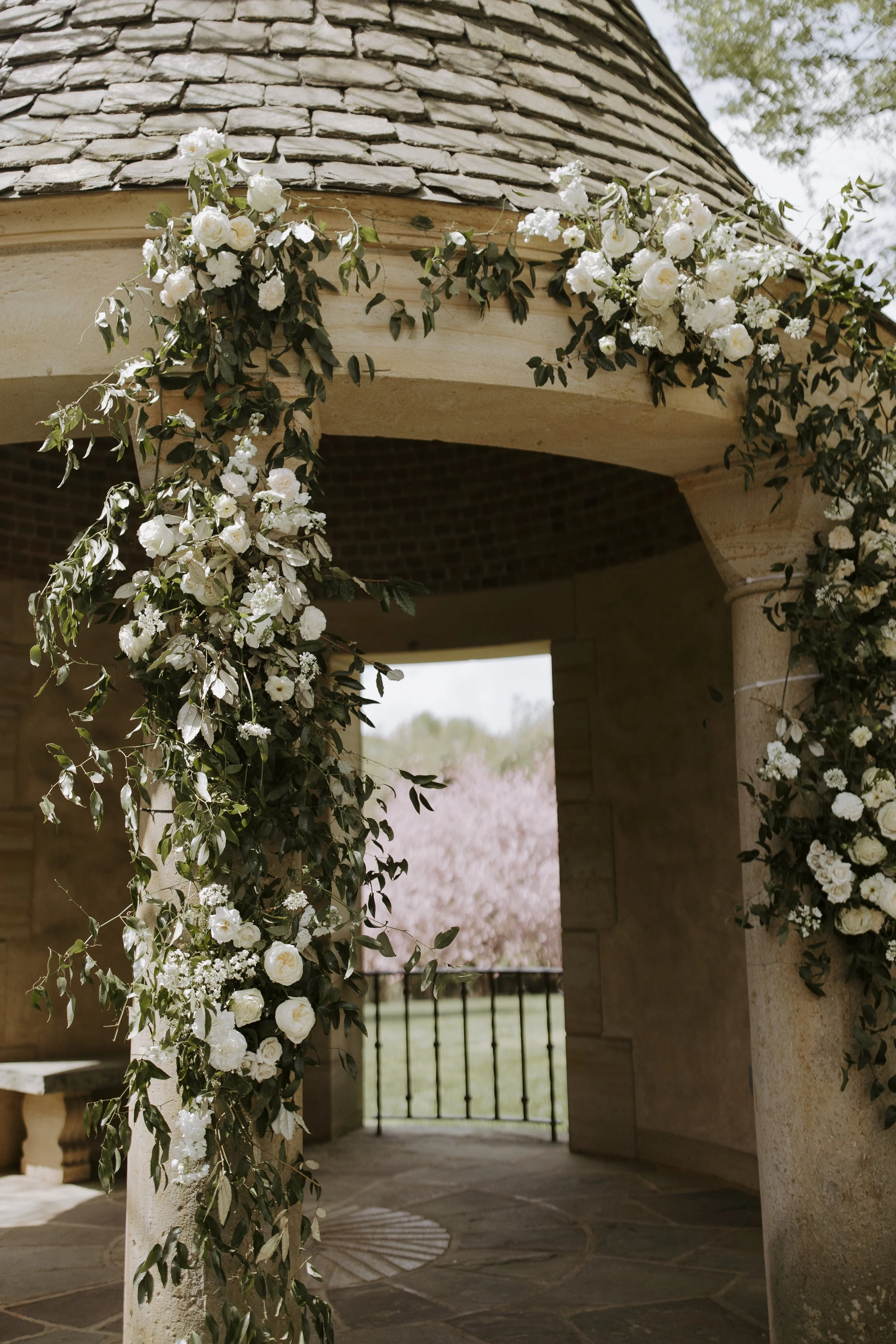 Decorated wedding arch with white flowers and green foliage, with a view of blooming trees in the background.