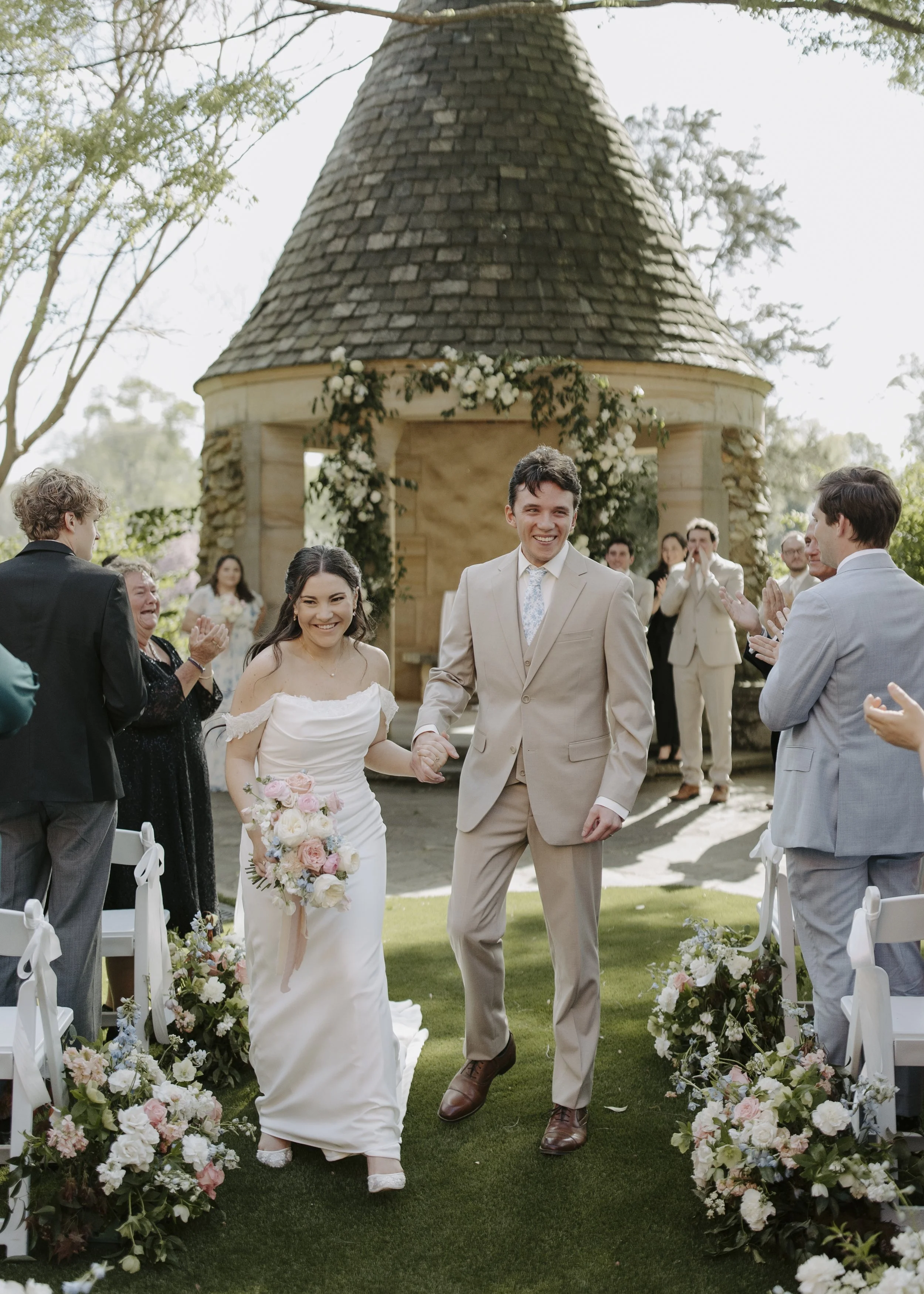 A newlywed couple walking down the aisle, holding hands and smiling. The bride is holding a bouquet of pink and white roses, and guests are clapping outdoors under a floral arch.