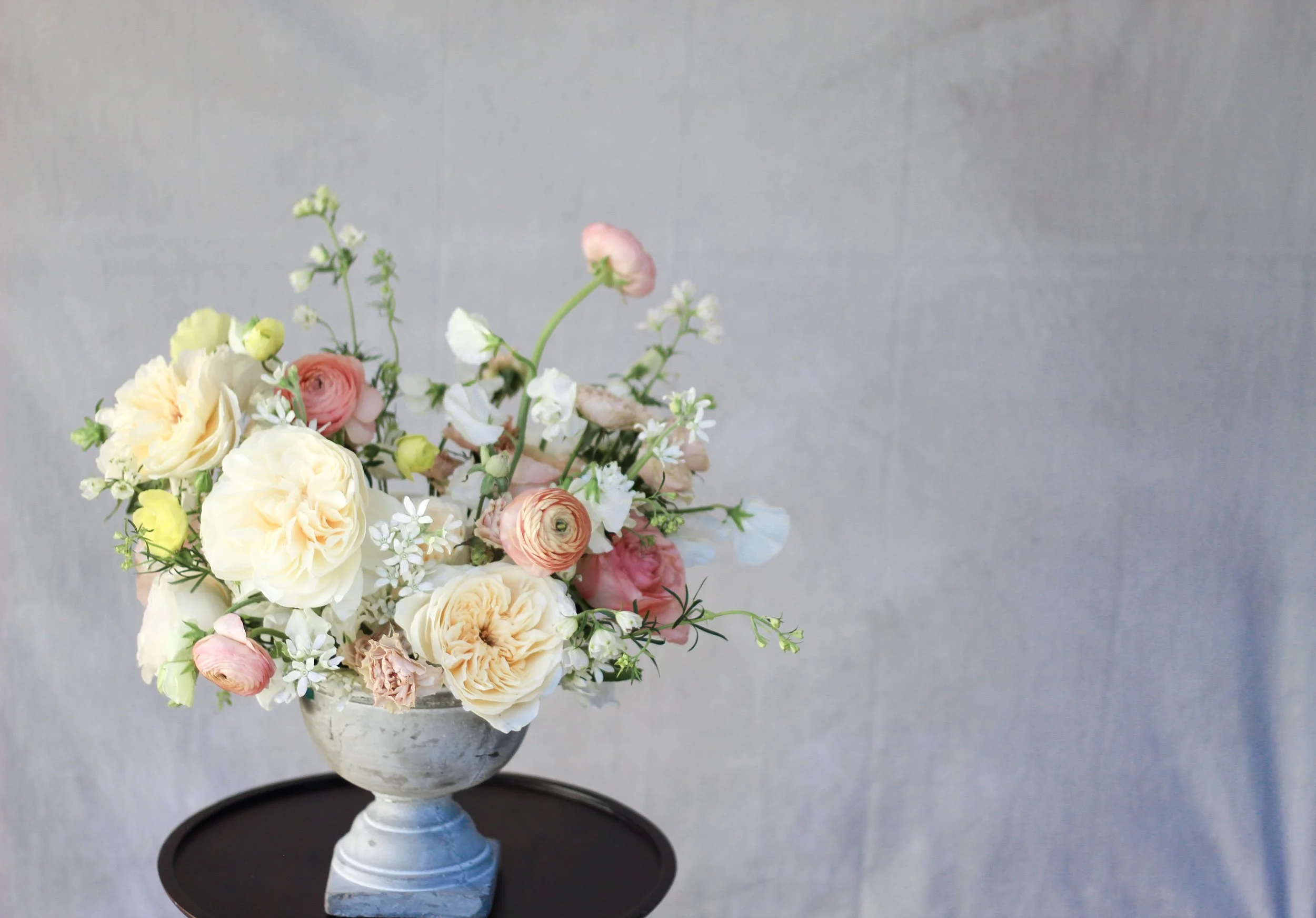 A bouquet of various white, peach, and pink flowers in a gray ceramic vase on a black round table against a plain gray backdrop.