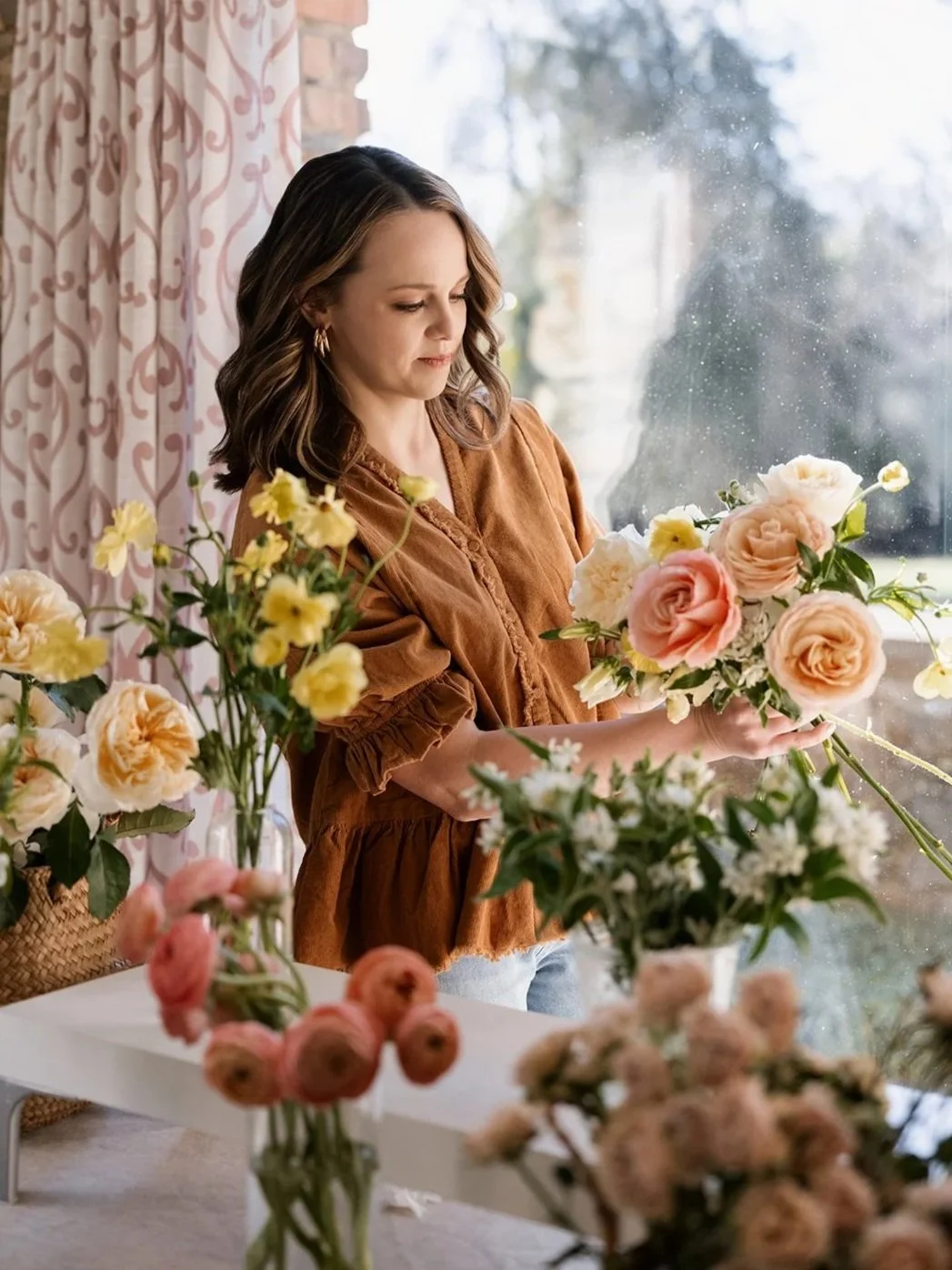 A woman arranging flowers indoors near a window with pink curtains.