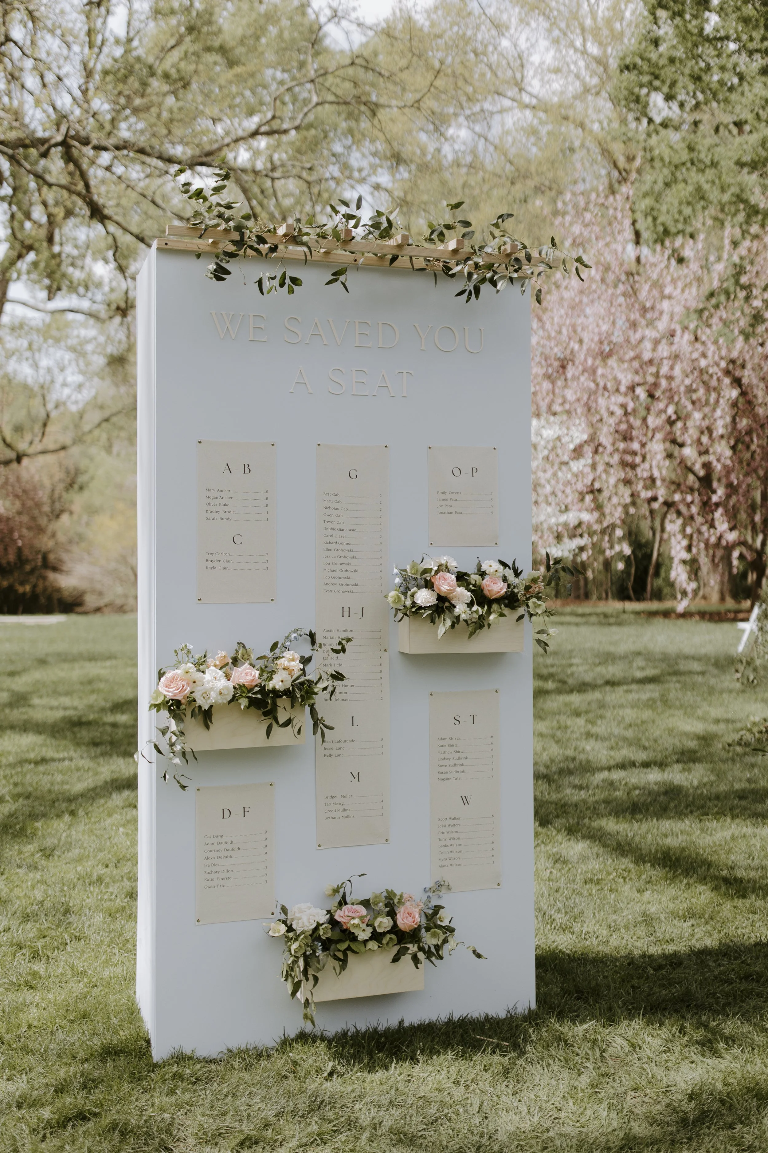 A seating chart board at an outdoor wedding, decorated with pink and white flowers and greenery, with trees and blooming flowers in the background.