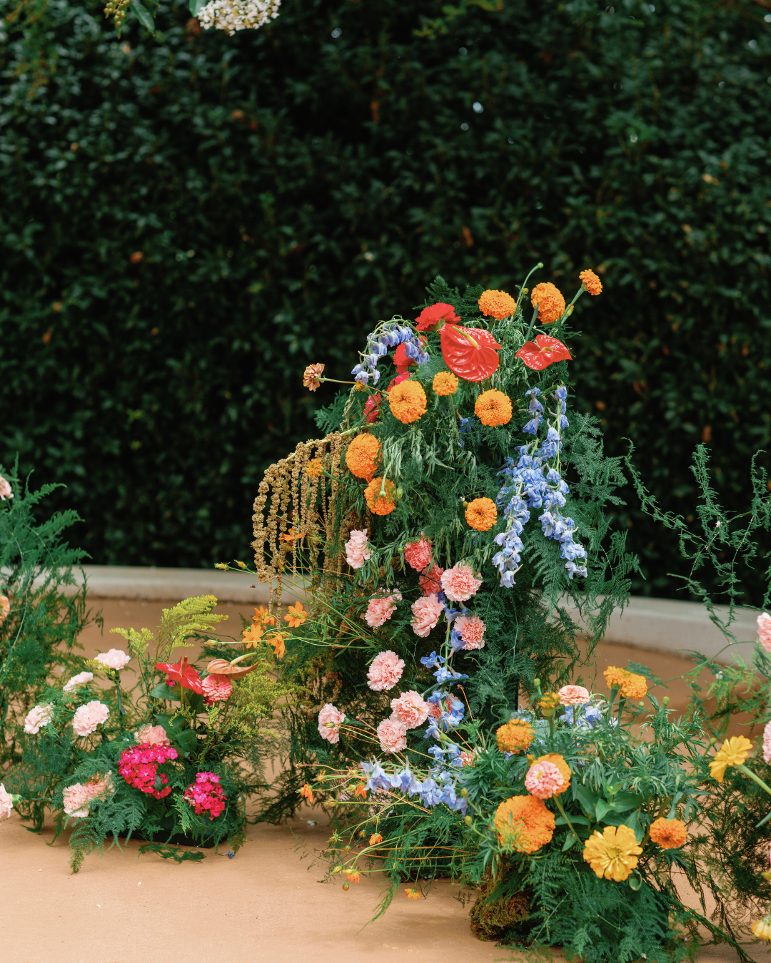 Colorful flower arrangement with orange marigolds, pink carnations, purple irises, red anthuriums, and yellow flowers against a dark green leafy background.