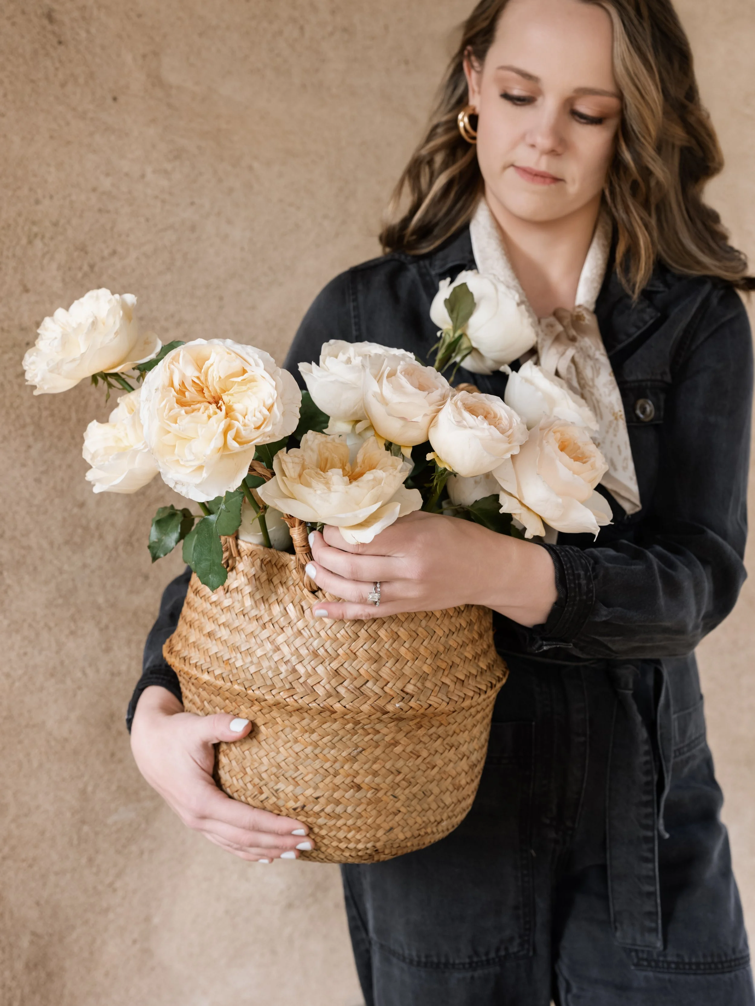 A woman holding a wicker basket filled with white and peach roses, standing against a beige textured wall.