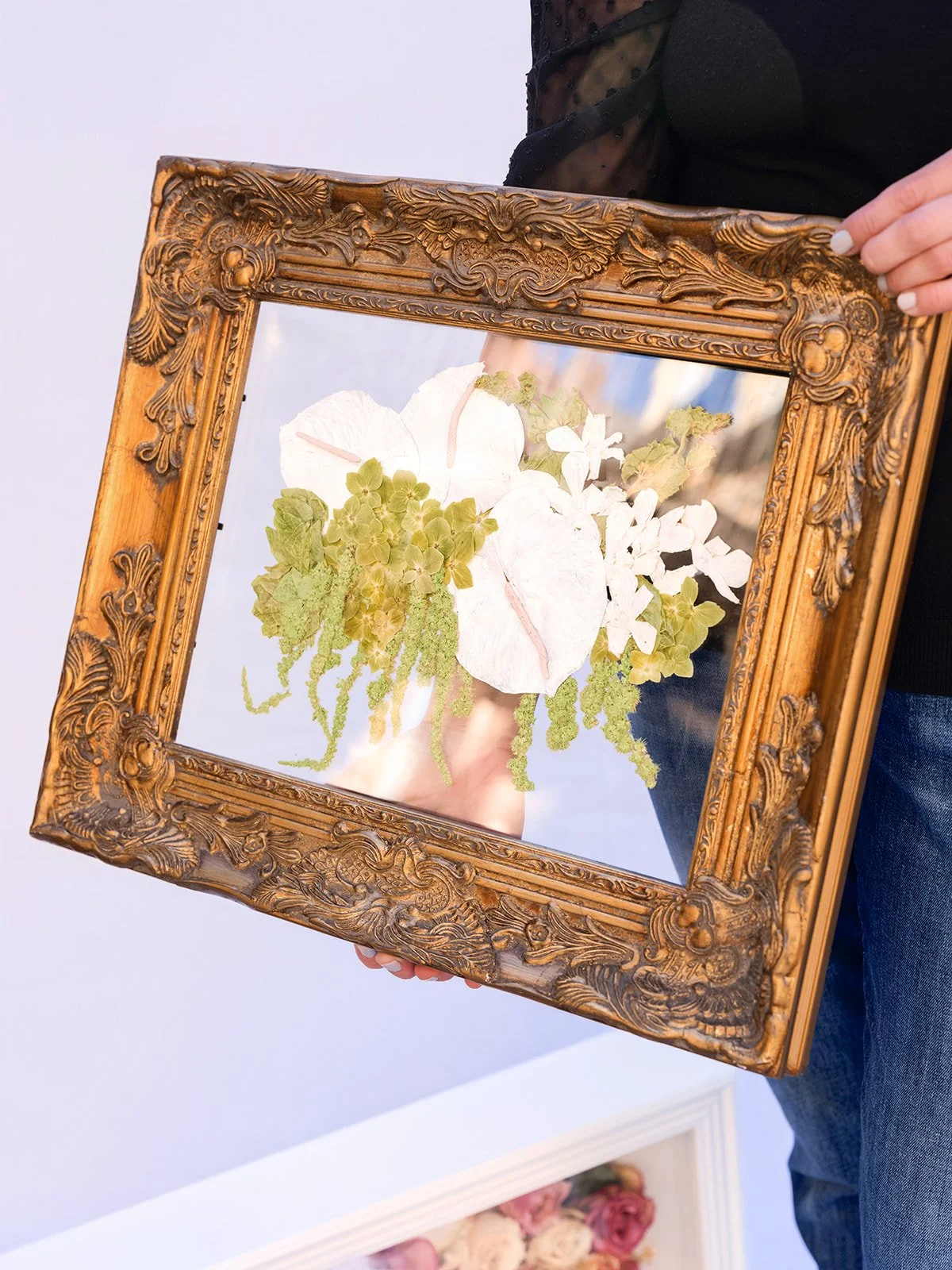 Person holding an ornate wooden framed mirror with dried flowers pressed against the glass, reflecting a blurry background.