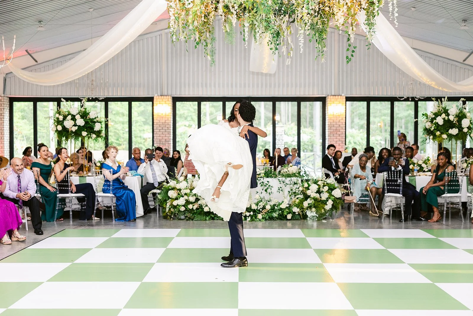 A bride and groom sharing a dance during their wedding reception in a decorated hall with large windows, white drapes, and floral arrangements.