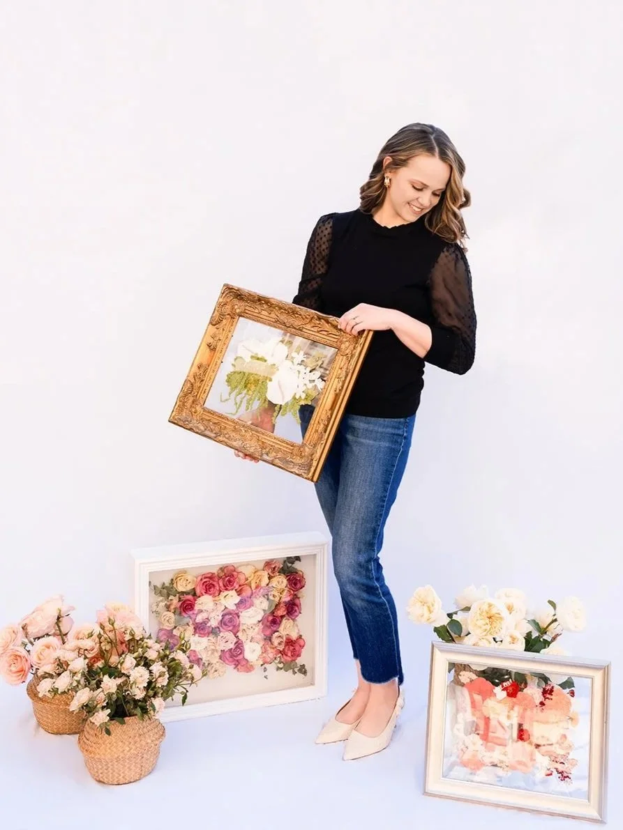 A woman holding a framed painting of white flowers, surrounded by additional floral artwork and bouquets of flowers on the floor.