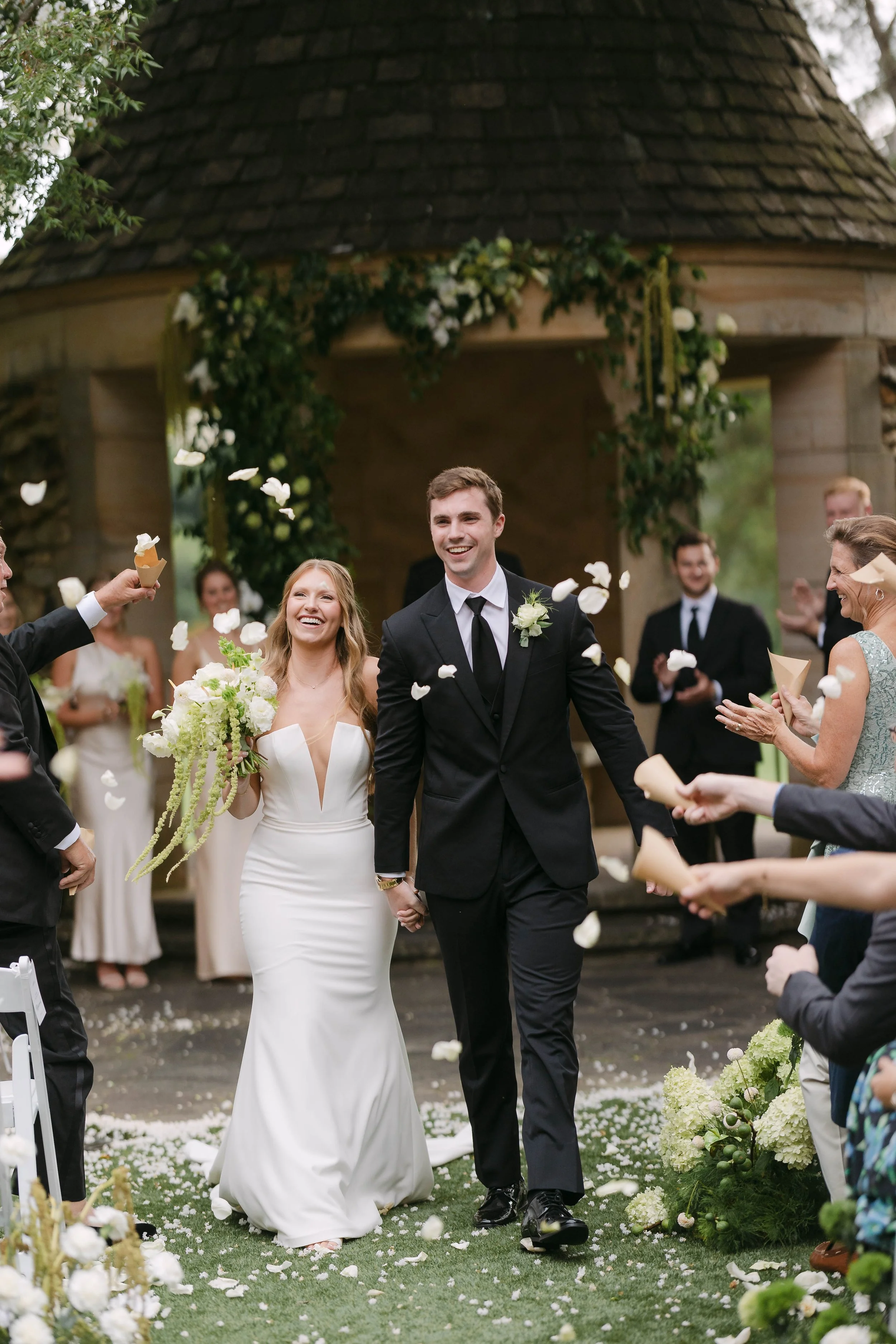Bride and groom walking hand in hand through guests throwing flower petals at an outdoor wedding ceremony.