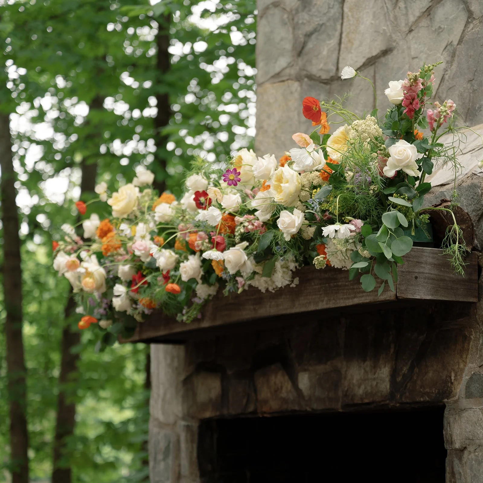 Colorful bouquet of flowers on a wooden mantel above a stone fireplace in a wooded setting.