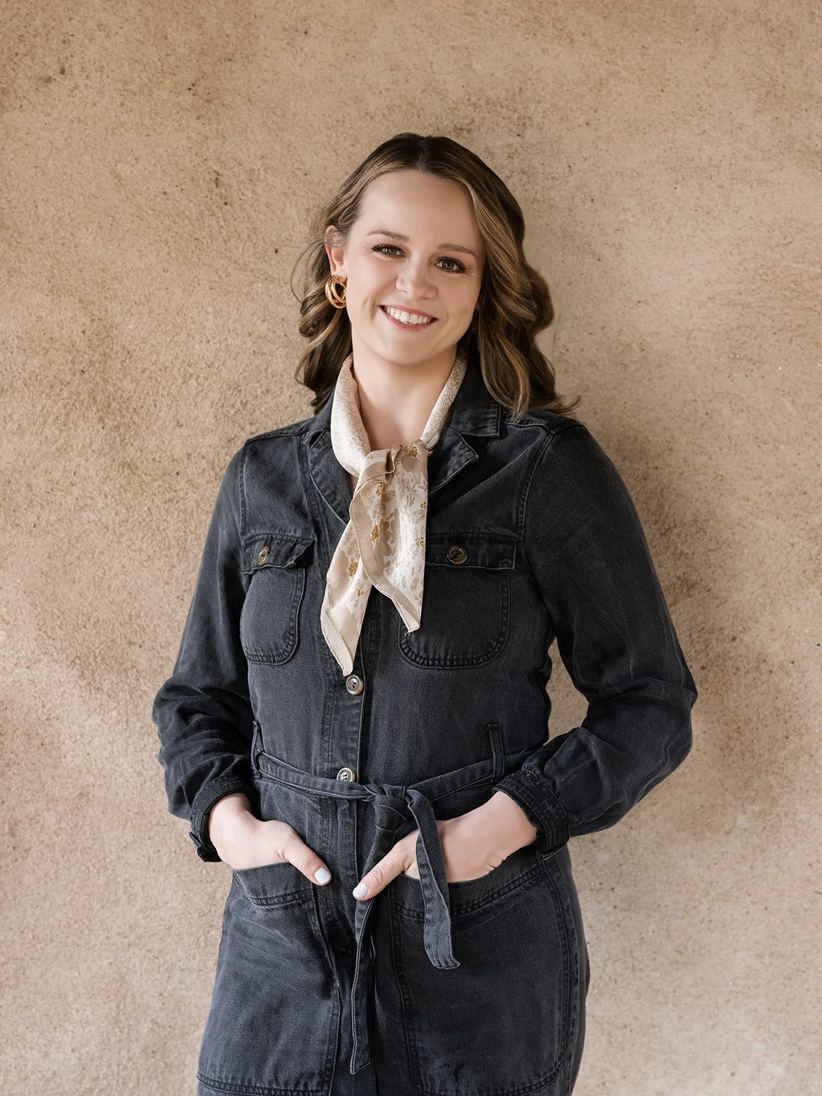 A woman with wavy brown hair, wearing hoop earrings, a beige silk scarf, and a dark denim jacket and matching pants, standing against a beige textured background and smiling at the camera.