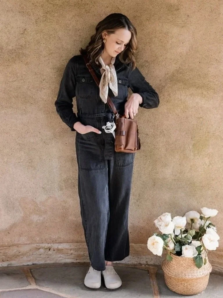 Woman wearing a black denim jumpsuit, light-colored scarf, white shoes, and carrying a brown crossbody bag, standing next to a basket of white flowers against a beige wall.