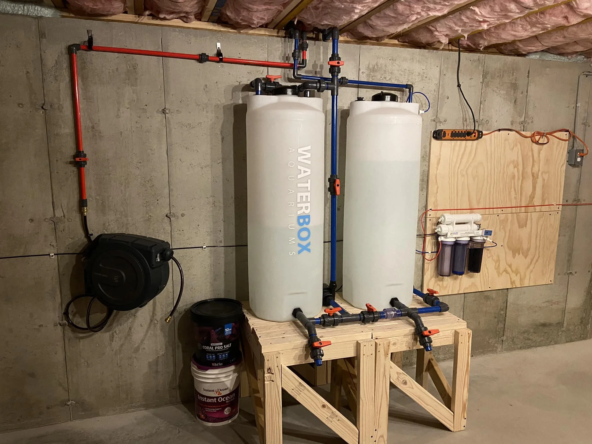 Water filtration system installed on a concrete wall with two large white tanks, a control panel, and pipes, set on a wooden stand, with salt buckets and a black hose reel nearby.