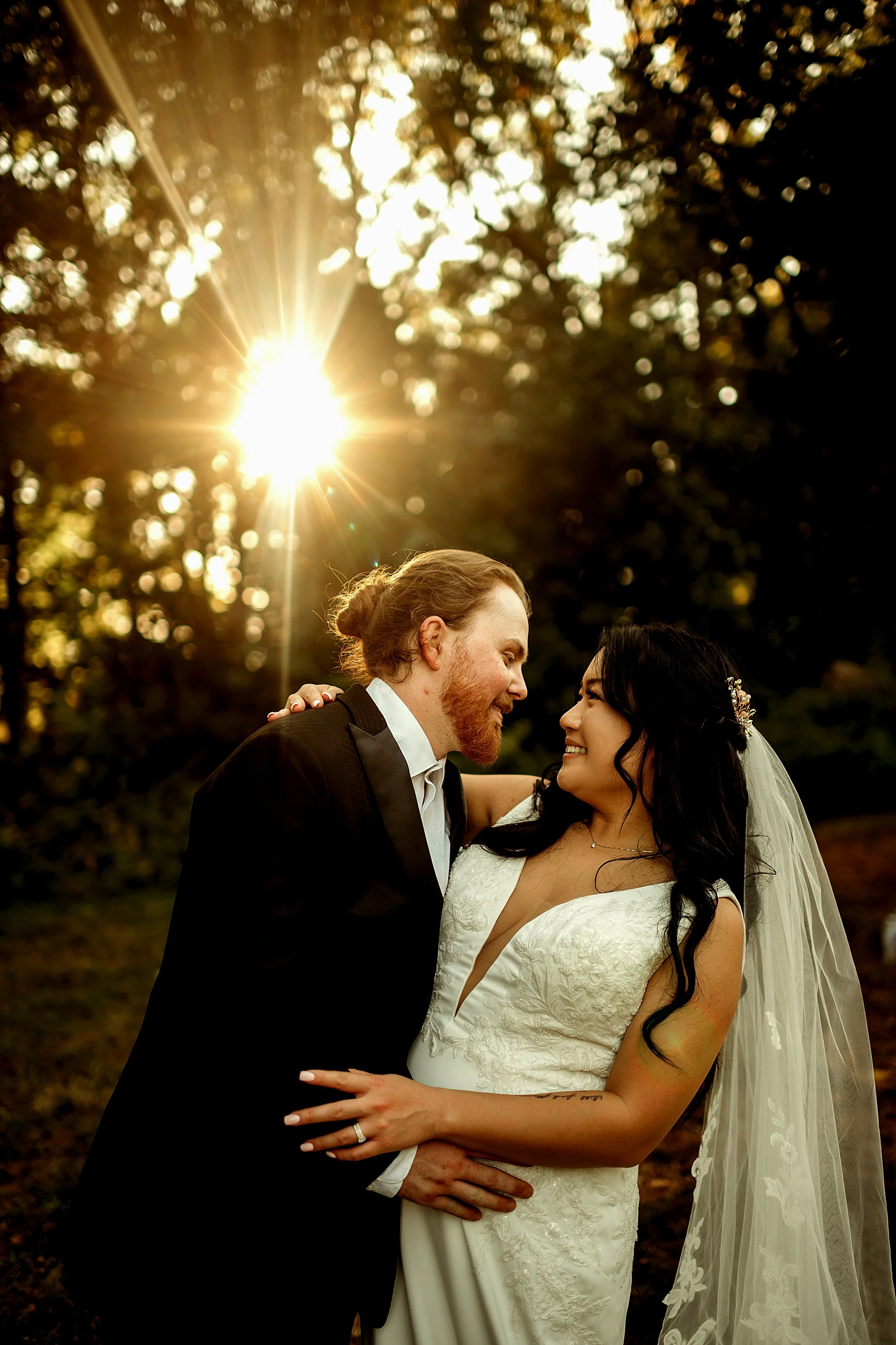 A wedding couple standing outdoors during sunset, sharing a joyful moment and smiling at each other, with trees and sunlight in the background.