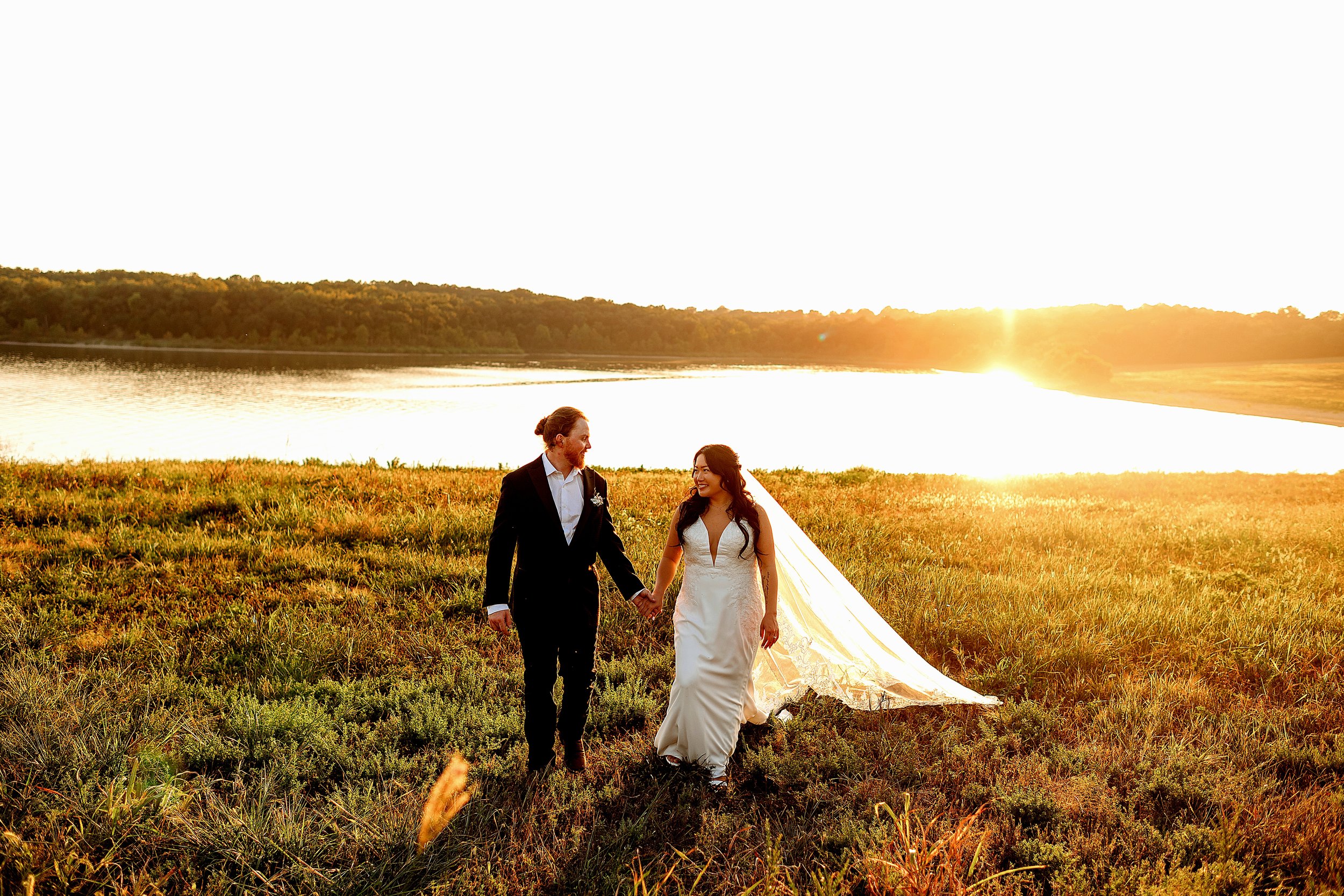 A bride and groom holding hands and walking on grass near a lake at sunset.