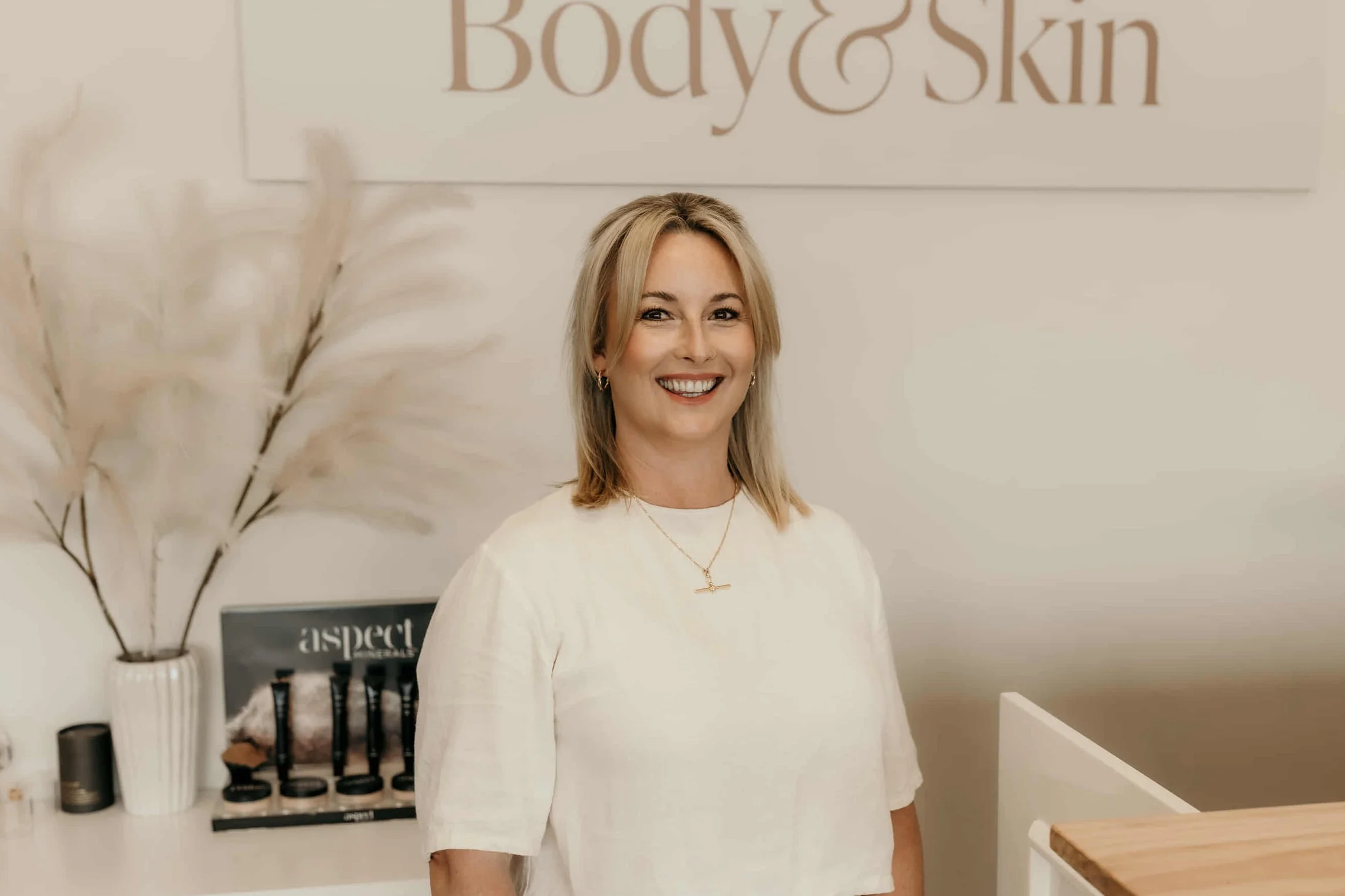 A smiling woman with blonde hair standing in a skincare or beauty salon, with a sign that reads 'Body & Skin' behind her, decorative pampas grass in a vase, and beauty products on a white counter.