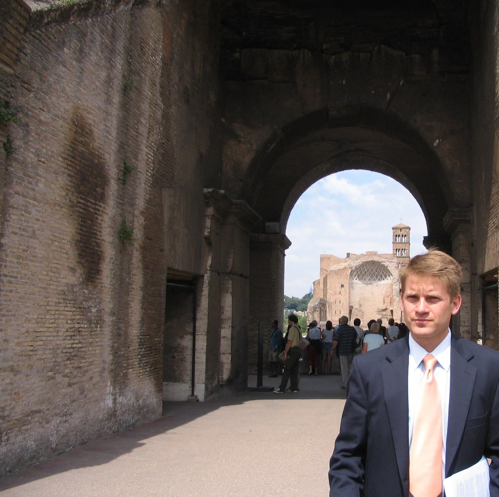 A man in a dark suit and light-colored tie stands in front of an ancient stone archway, with ruins and a domed structure visible in the background and a group of tourists walking nearby.