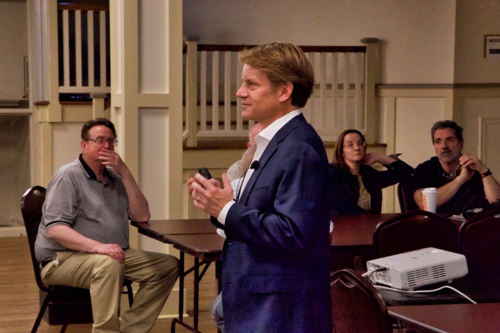 A man in a blue suit giving a presentation to an audience in a conference room, with three seated attendees listening.
