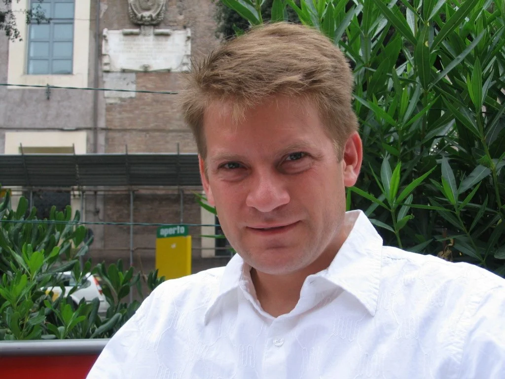 A man with light brown hair wearing a white shirt sitting outdoors with green plants and a building in the background.