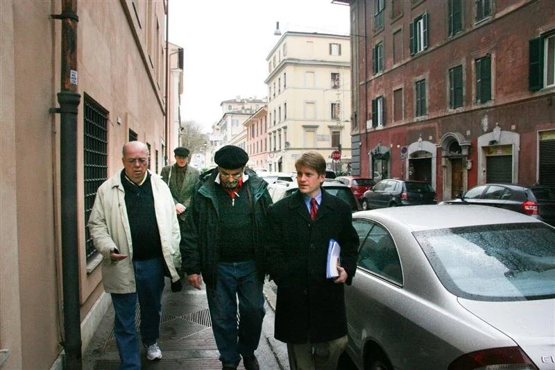 Four men walking on a city sidewalk, with parked cars and multi-story buildings in the background. One man is holding a notebook or folder.