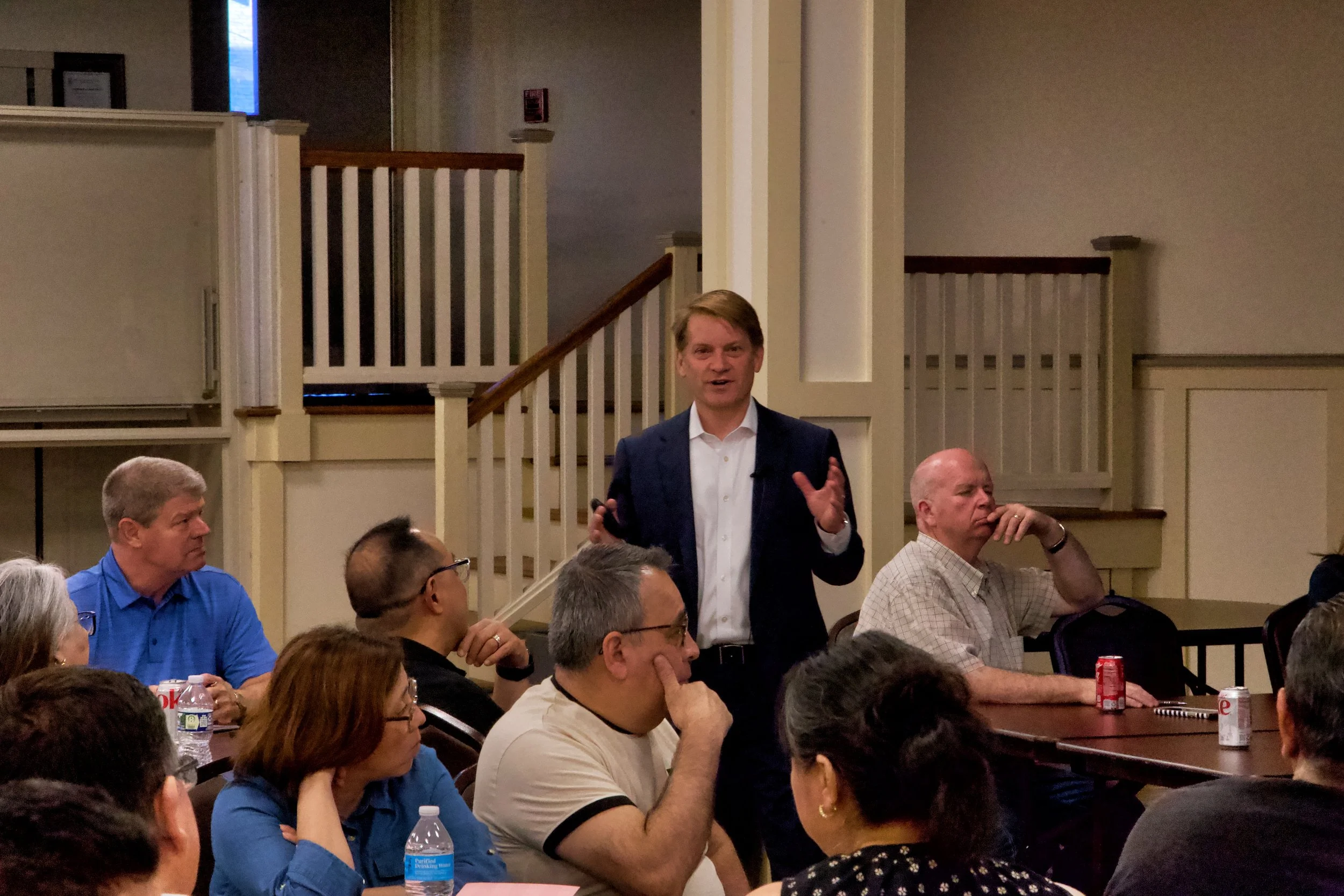 A man in a dark suit and white shirt giving a presentation to a group of people seated at tables in a conference room.