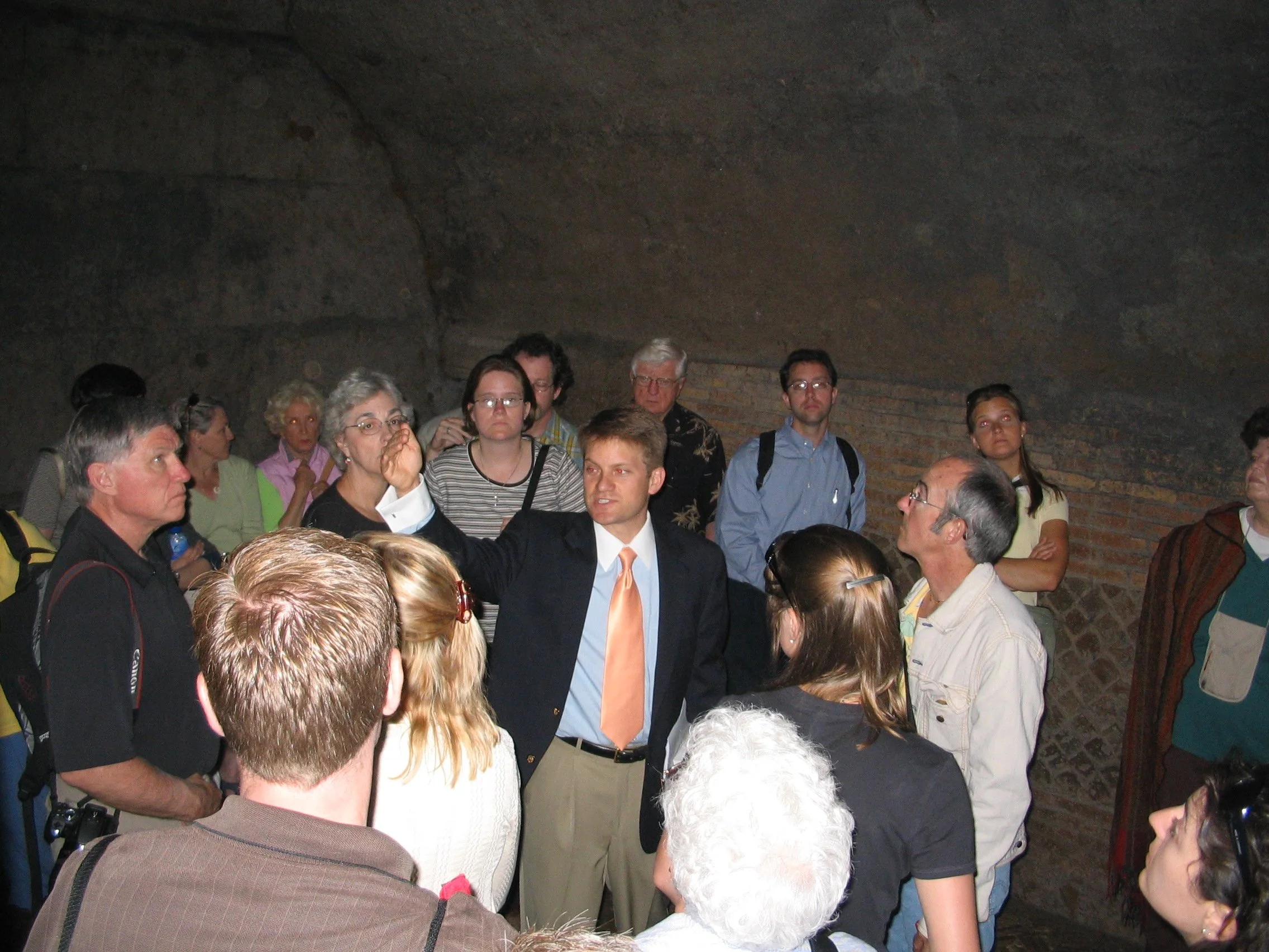Group of people gathered inside a cave or underground chamber, listening to a man in a suit and orange tie explaining something.