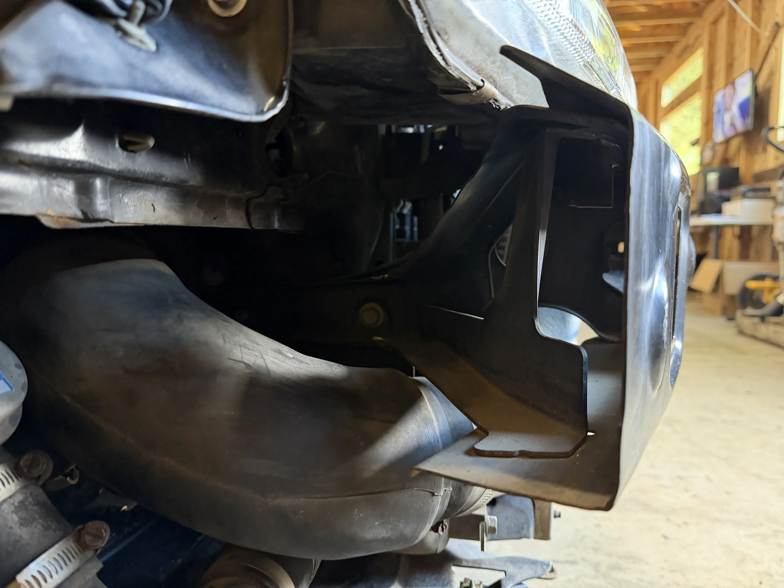 Close-up view of a car engine with a black metal bracket attached, in a garage with tools and a TV in the background.