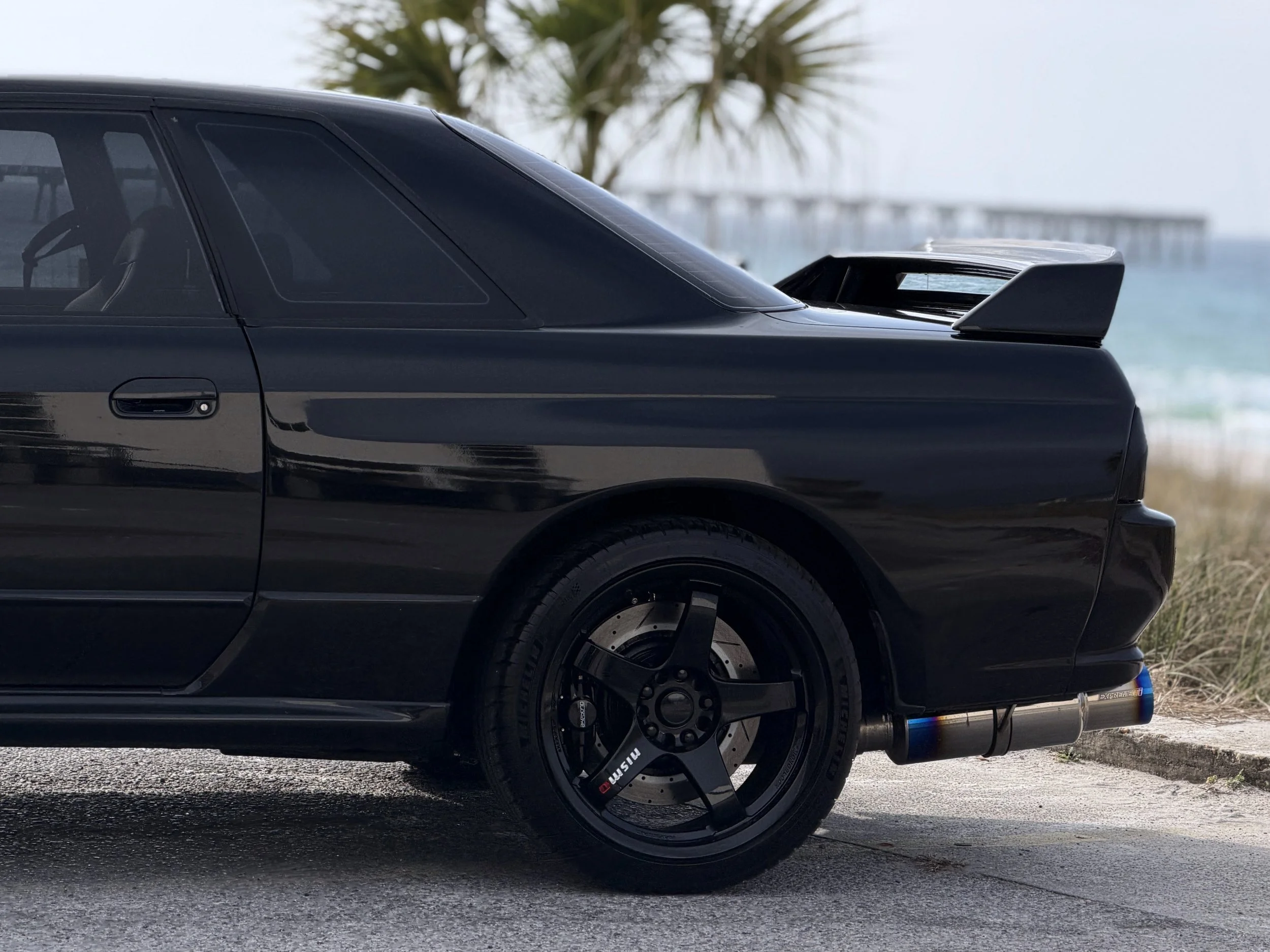Close-up of the rear side of a black sports car parked on a street near a body of water with a bridge in the background. The car features a large rear wing spoiler and performance wheels.