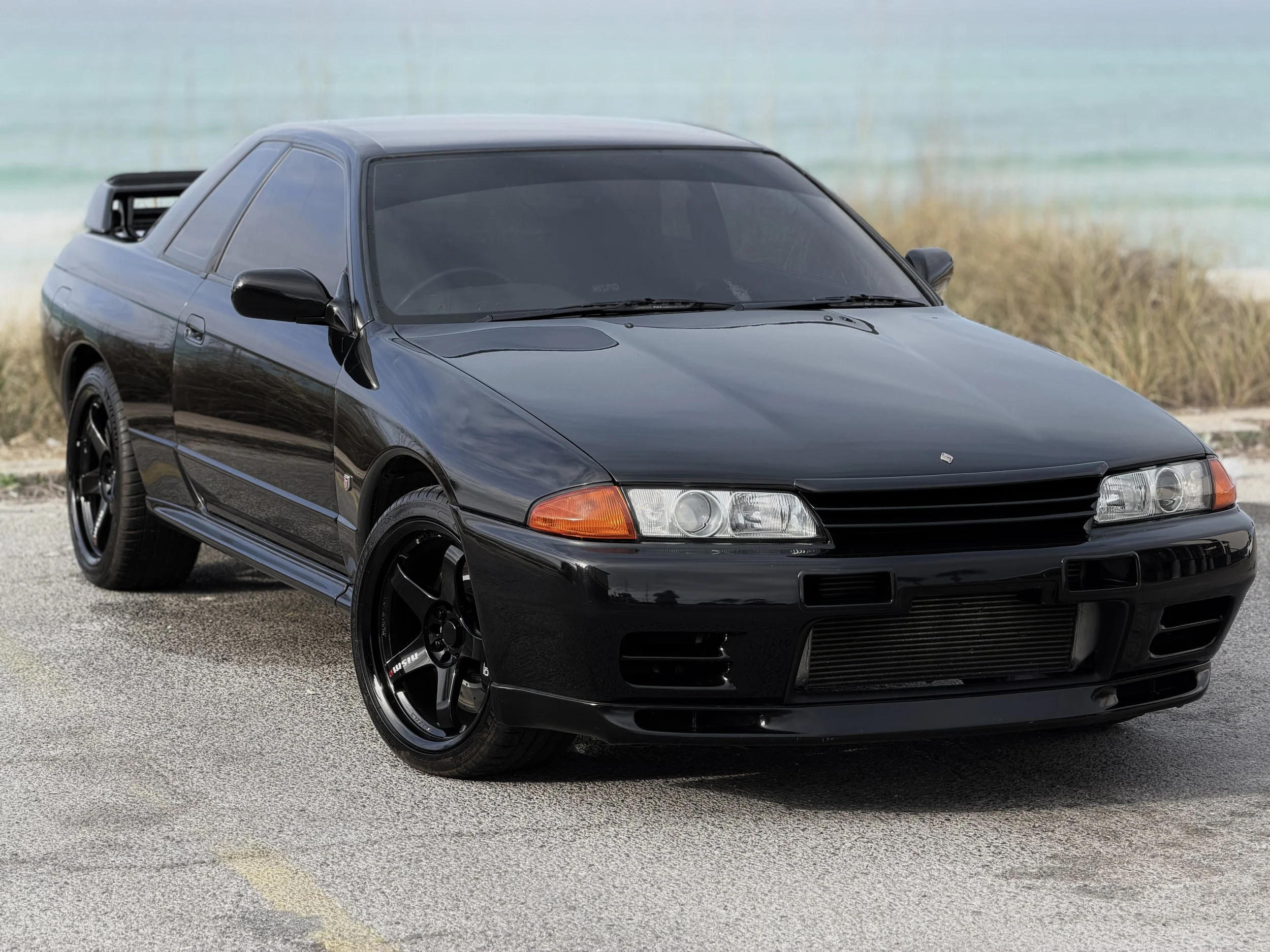 R32 NISSAN SKYLINE GTR FOR SALE
Black Nissan Skyline GTR parked near a beach with yellow guard lines on the pavement.