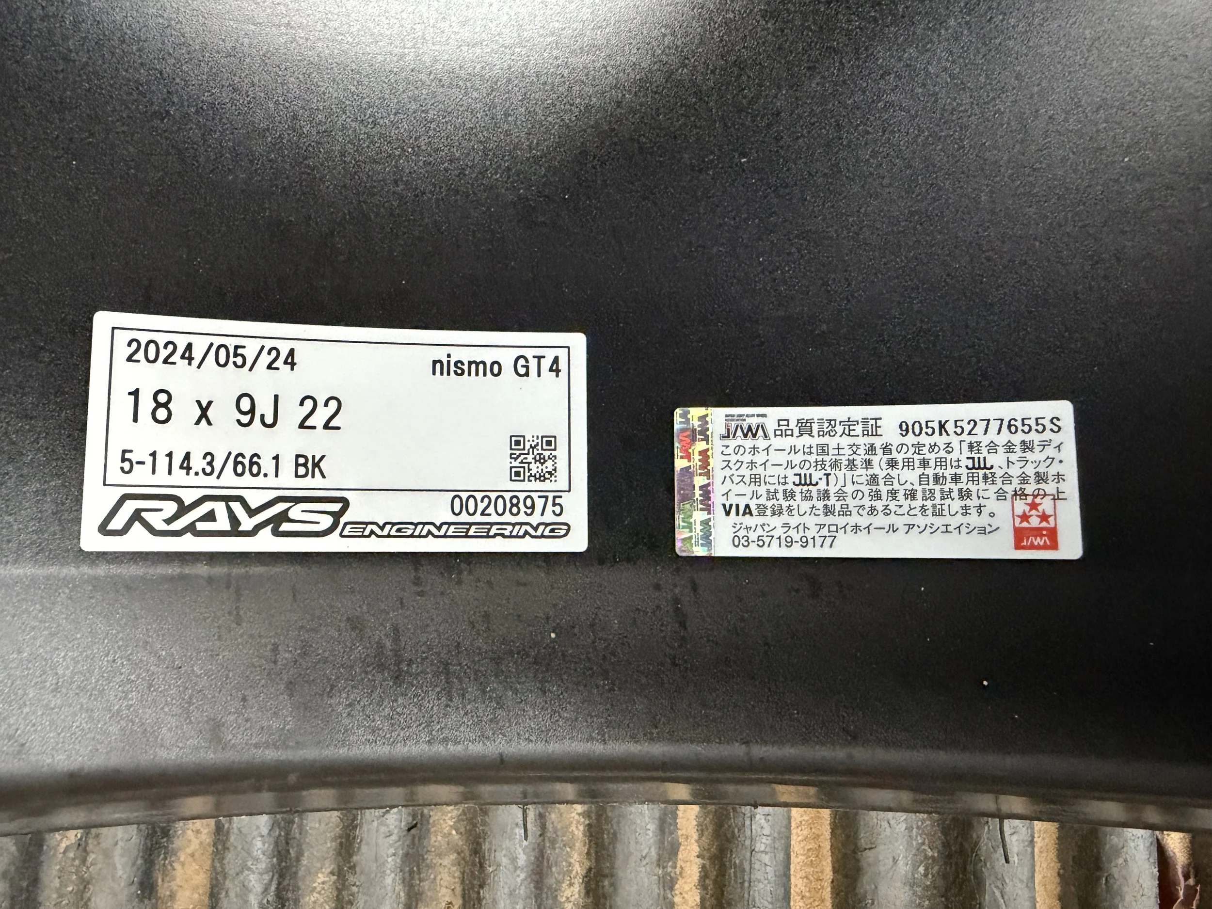 Close-up of two labels on a black surface. The left label is for a Nismo GT4 wheel, with specifications and a QR code. The right label is certification info in Japanese, including a red logo and additional specifications.