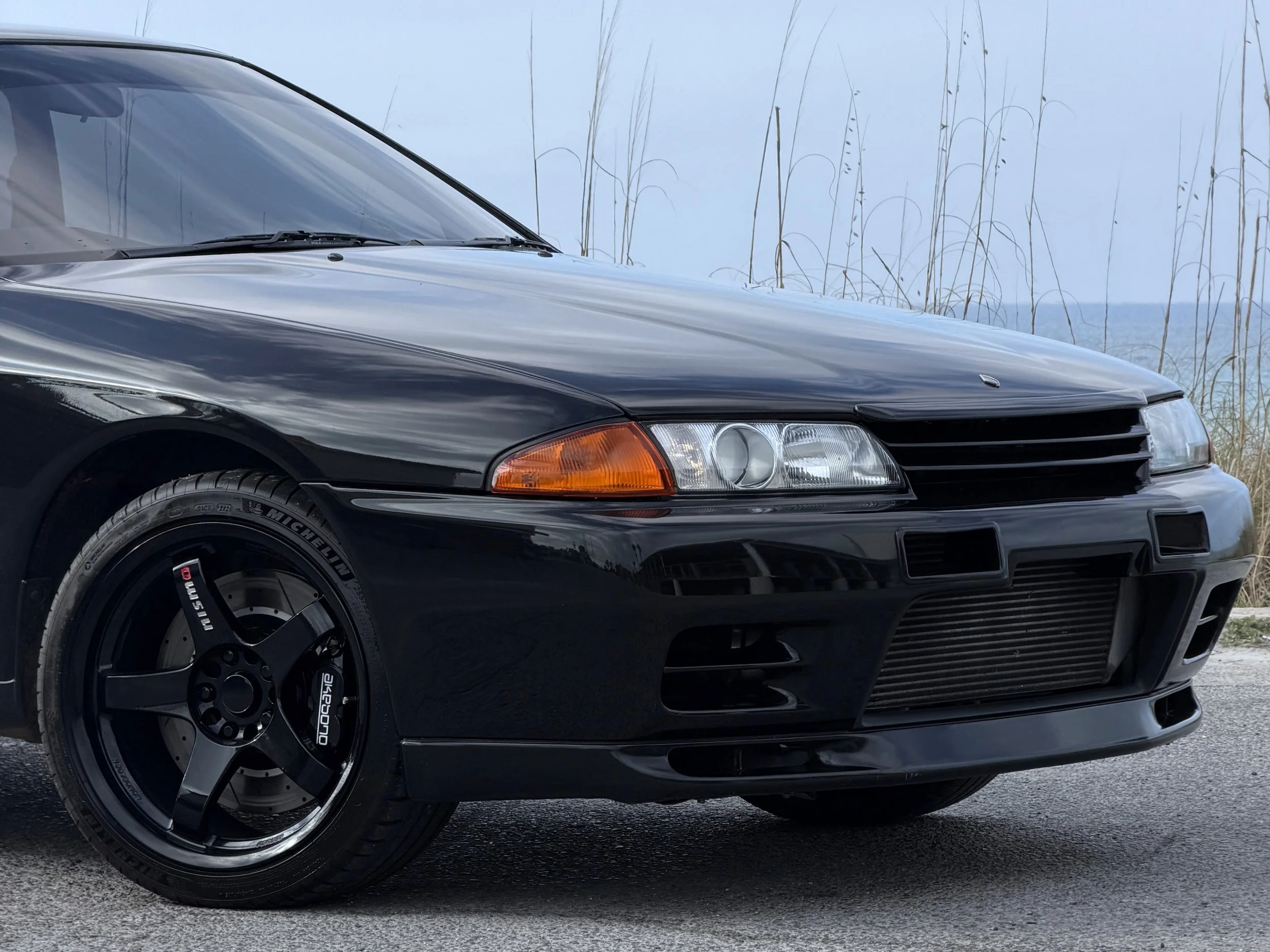 Close-up of the front section of a black sports car parked on a paved surface, with a background of tall, dry grass and a cloudy sky.
