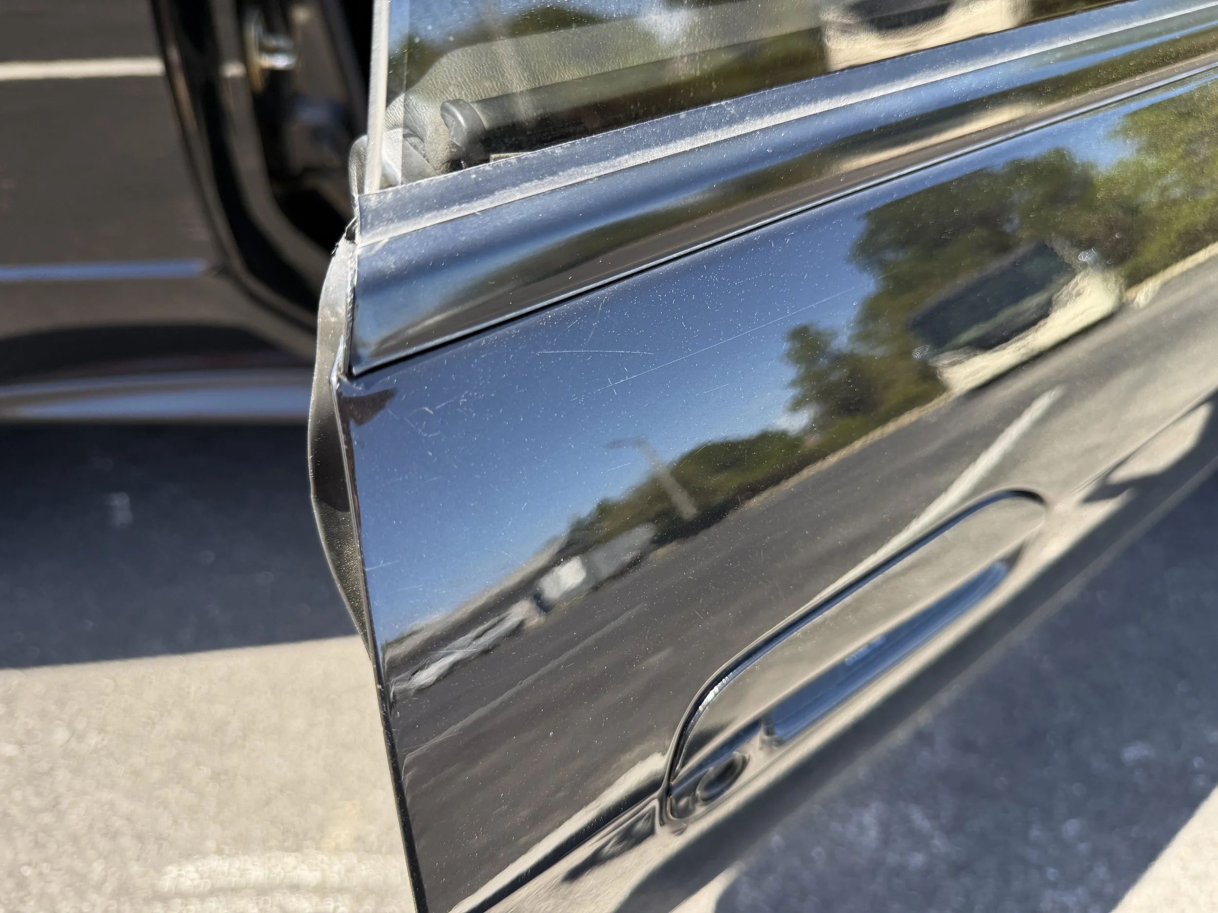 Close-up of a black car door with a reflective surface, showing reflections of trees and sky, and some minor surface scratches.
