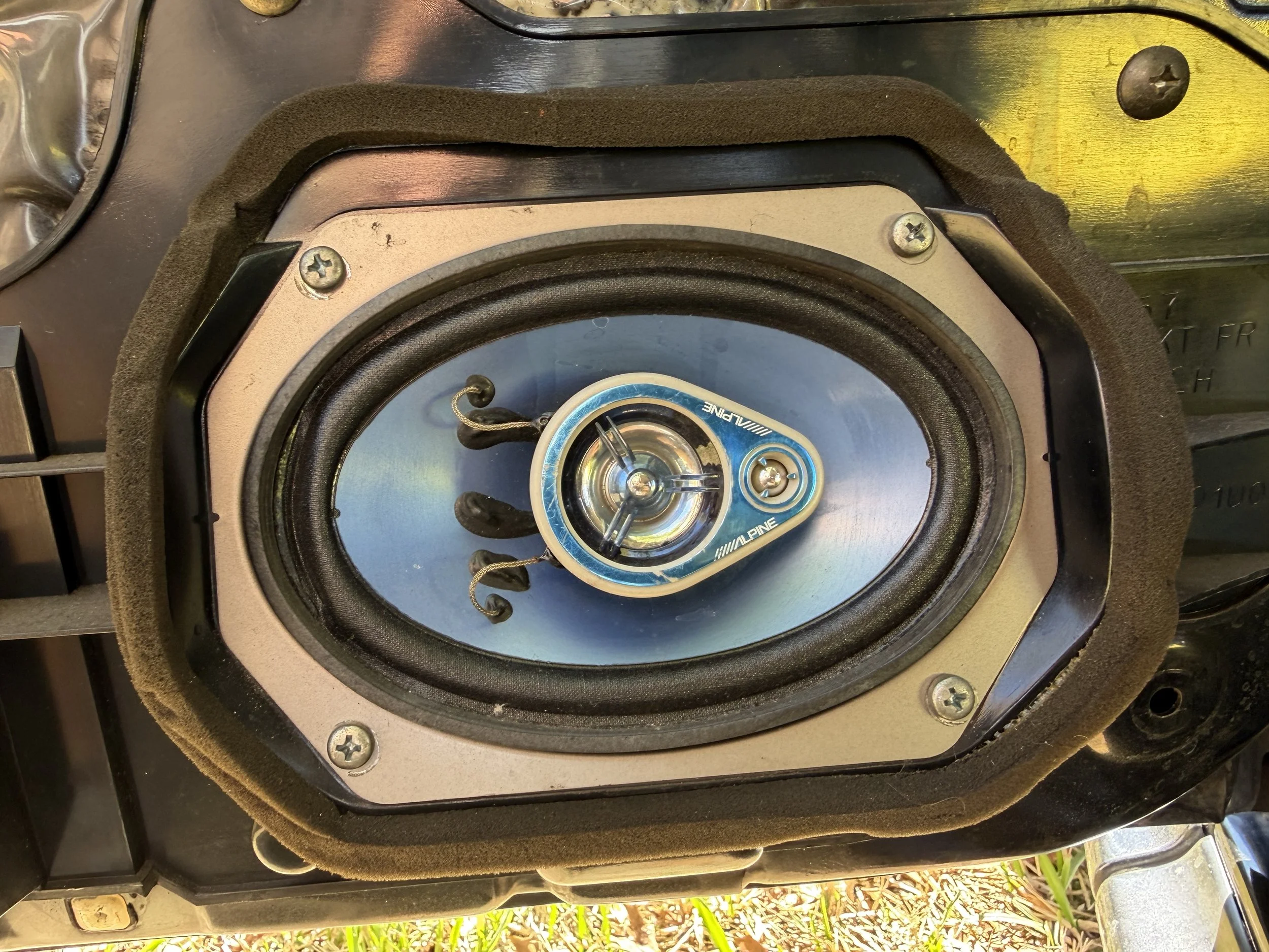 Close-up of a speaker with a blue and black Alpine component, mounted in a black speaker enclosure with screws and foam padding.