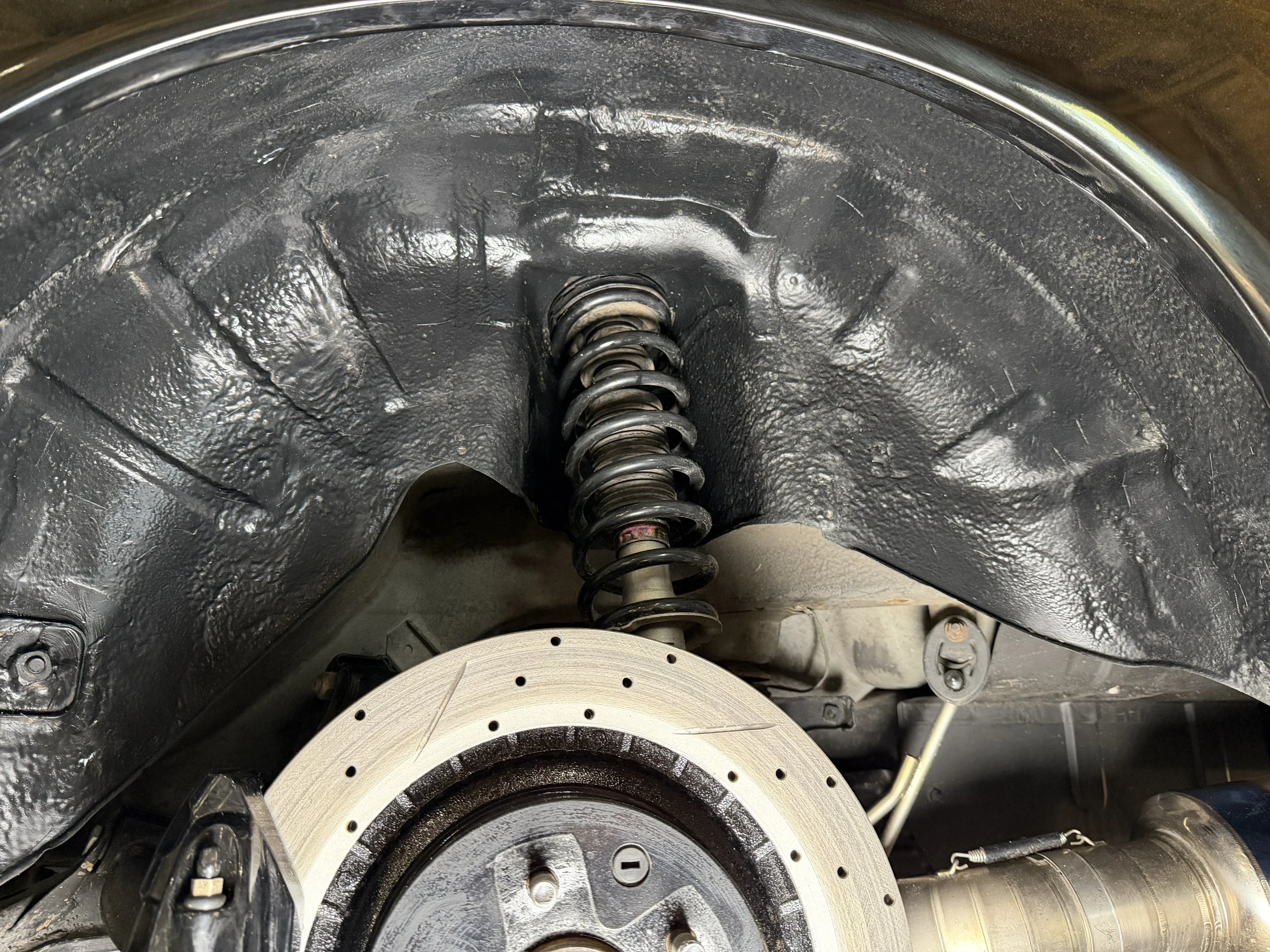 Close-up of a car's wheel well showing a black suspension spring, part of the brake rotor, and surrounding components.