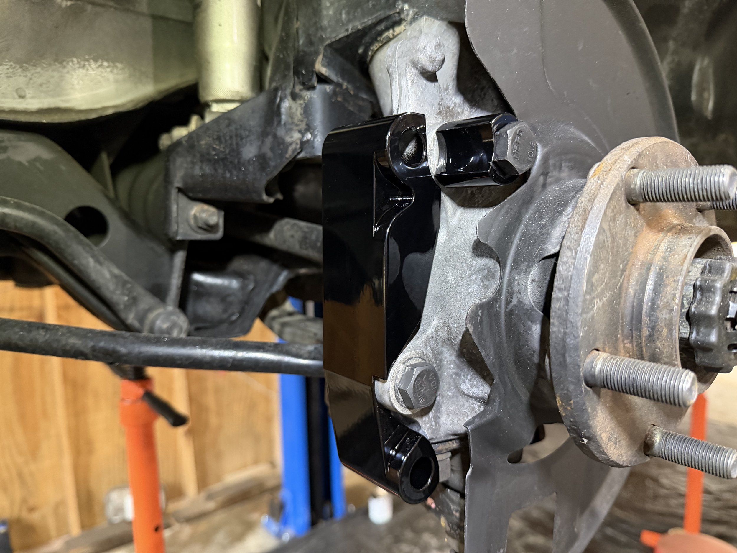 A black metal bracket attached to a vehicle's wheel hub assembly with bolts, next to rusty wheel studs and hub components.