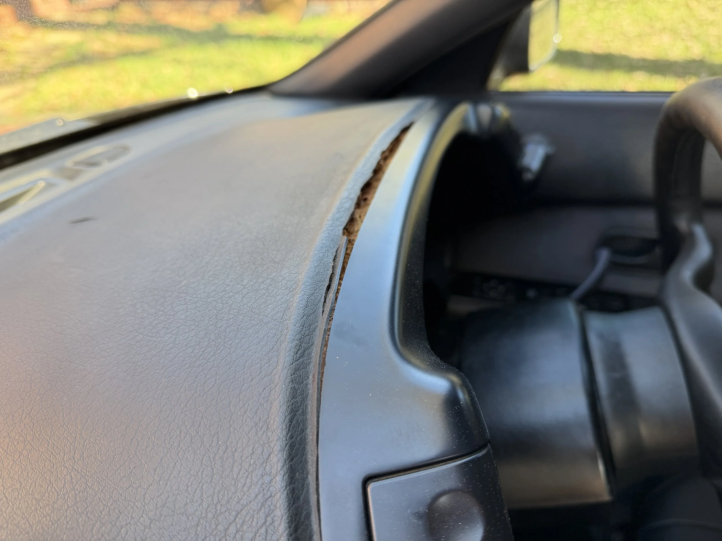Close-up of a car's dashboard with a crack and missing piece in the plastic near the windshield on a sunny day.